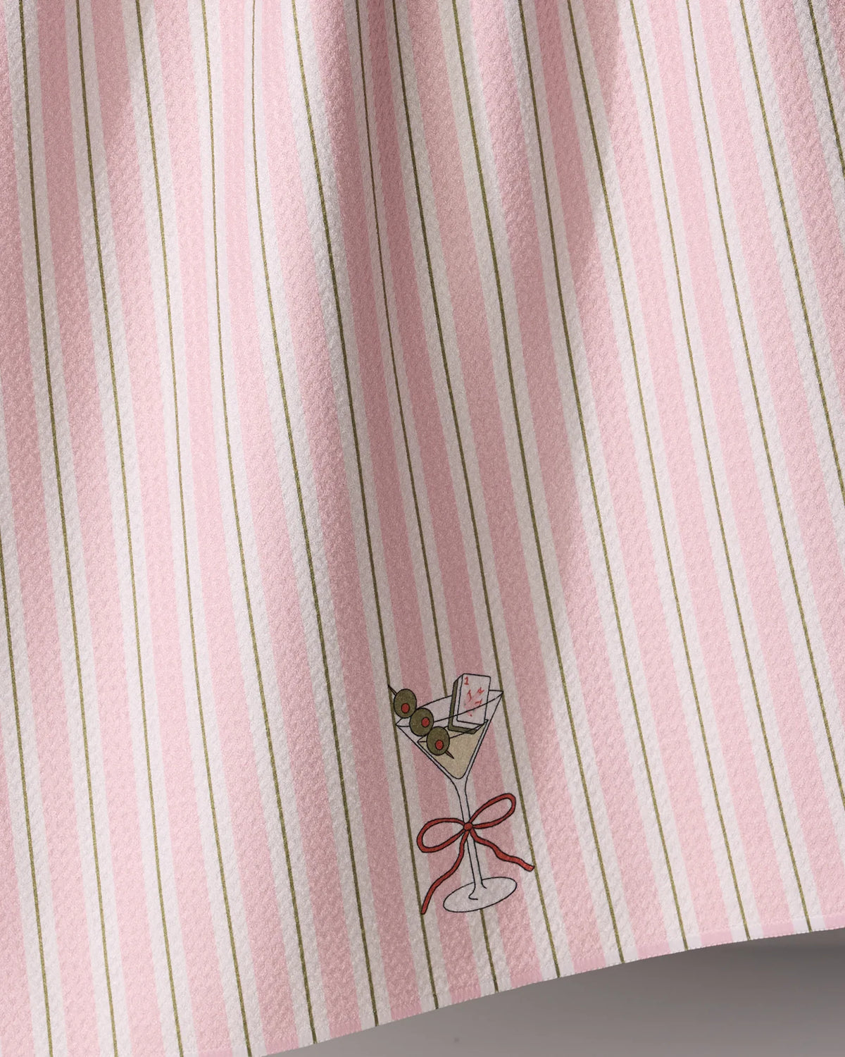 A Geometry Martini Stripe kitchen towel with thin vertical pink and green stripes, an illustration of a martini glass with olives and a playing card, and a red ribbon tie.