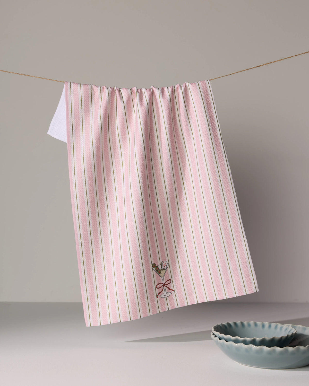 A Geometry Martini Stripe tea towel with pink and white stripes hanging on a line, featuring an embroidered martini glass with a red bow at the bottom.