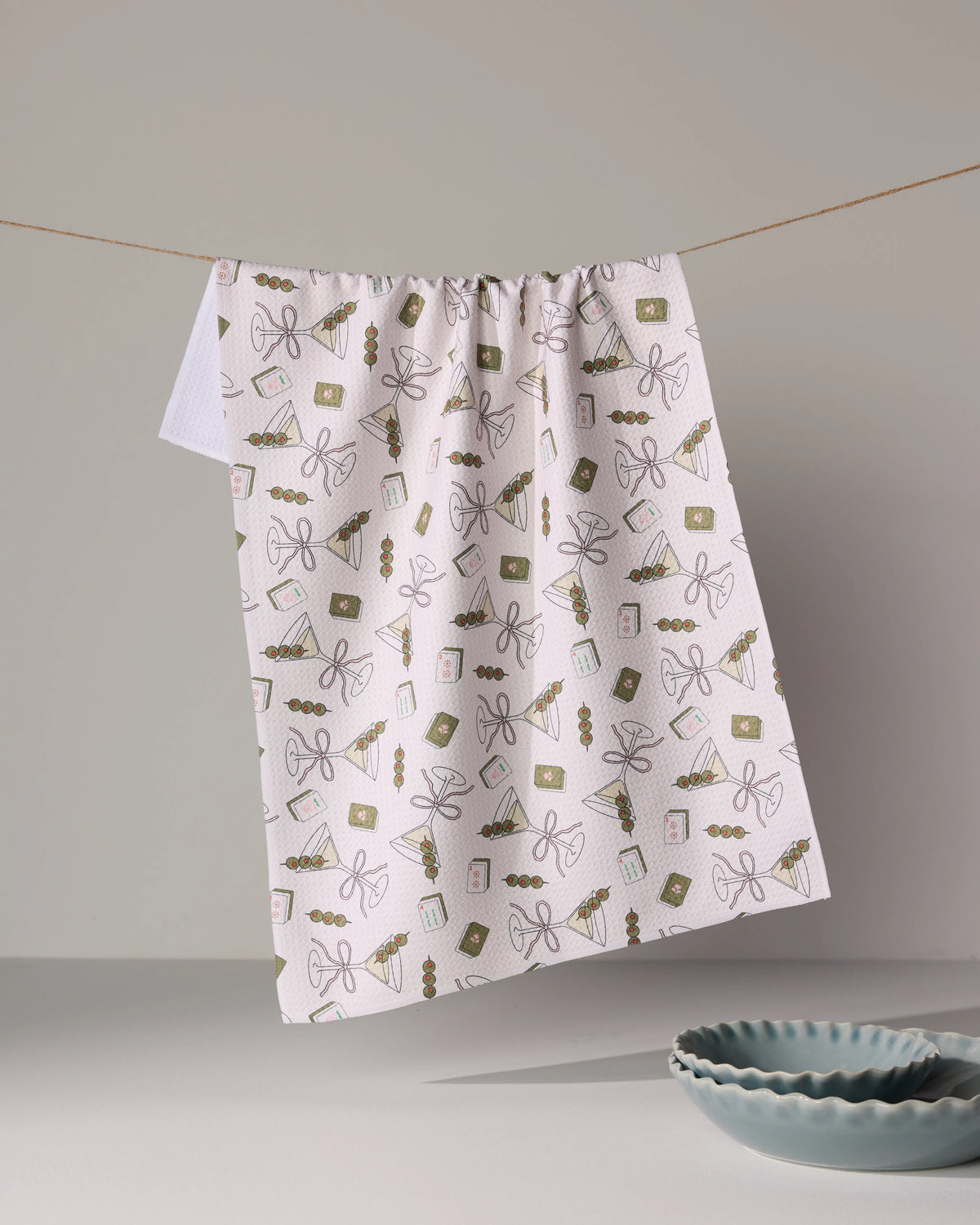 Geometry Martini Tiles ultra-absorbent white kitchen towel with a repeating pattern of martini glasses, olive picks, and small books hanging on a string above two blue bowls.