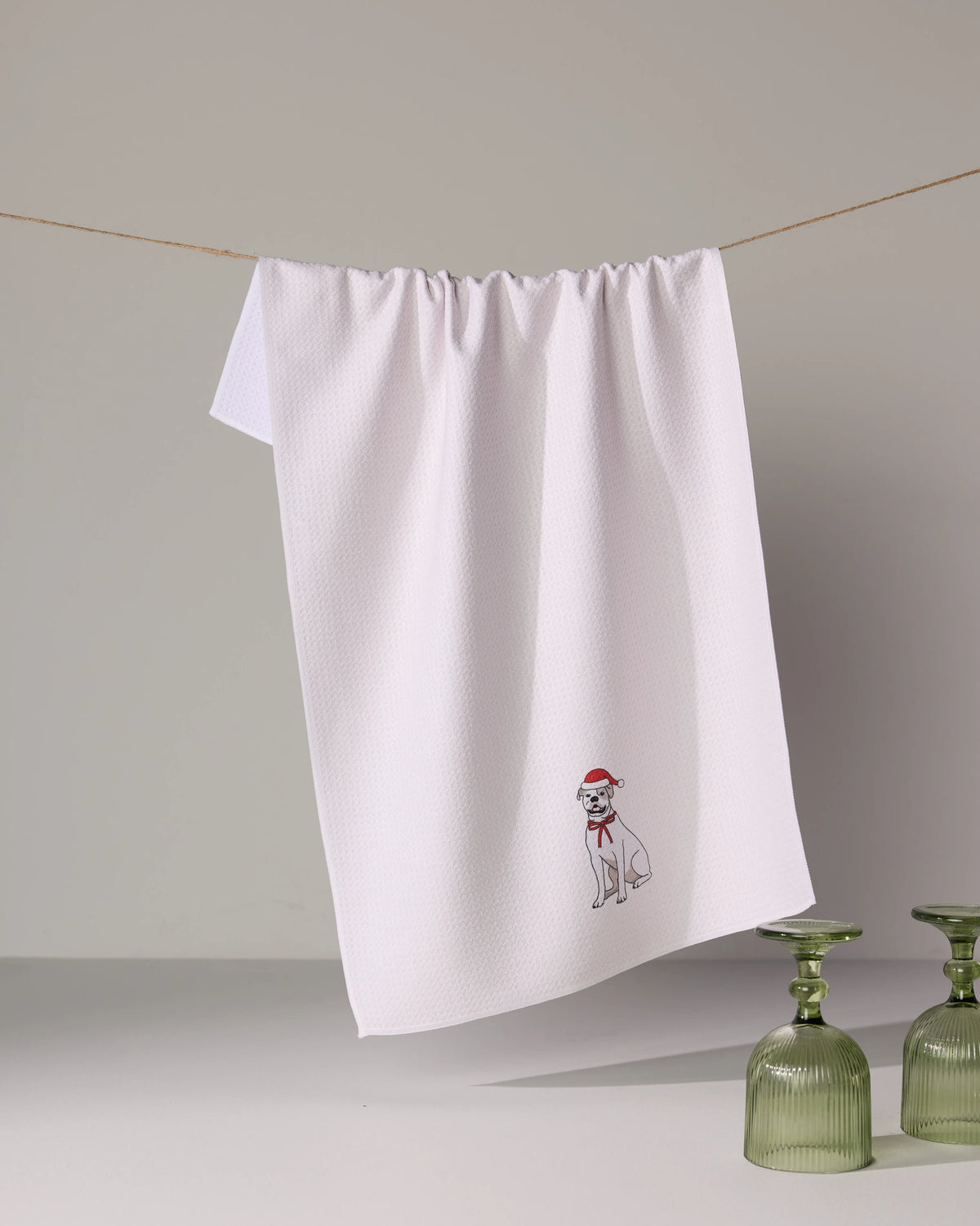 The Geometry Merry Bulldog, a white quick-drying tea towel with an embroidered dog in a red Santa hat and scarf, hangs above two upside-down green glass cups.