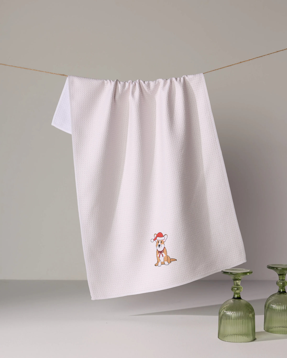 The Geometry Merry Corgi is a white tea towel featuring a small embroidered cartoon corgi in a red hat, hanging above two green glass cups. Made from quick-drying, recycled materials for daily use.