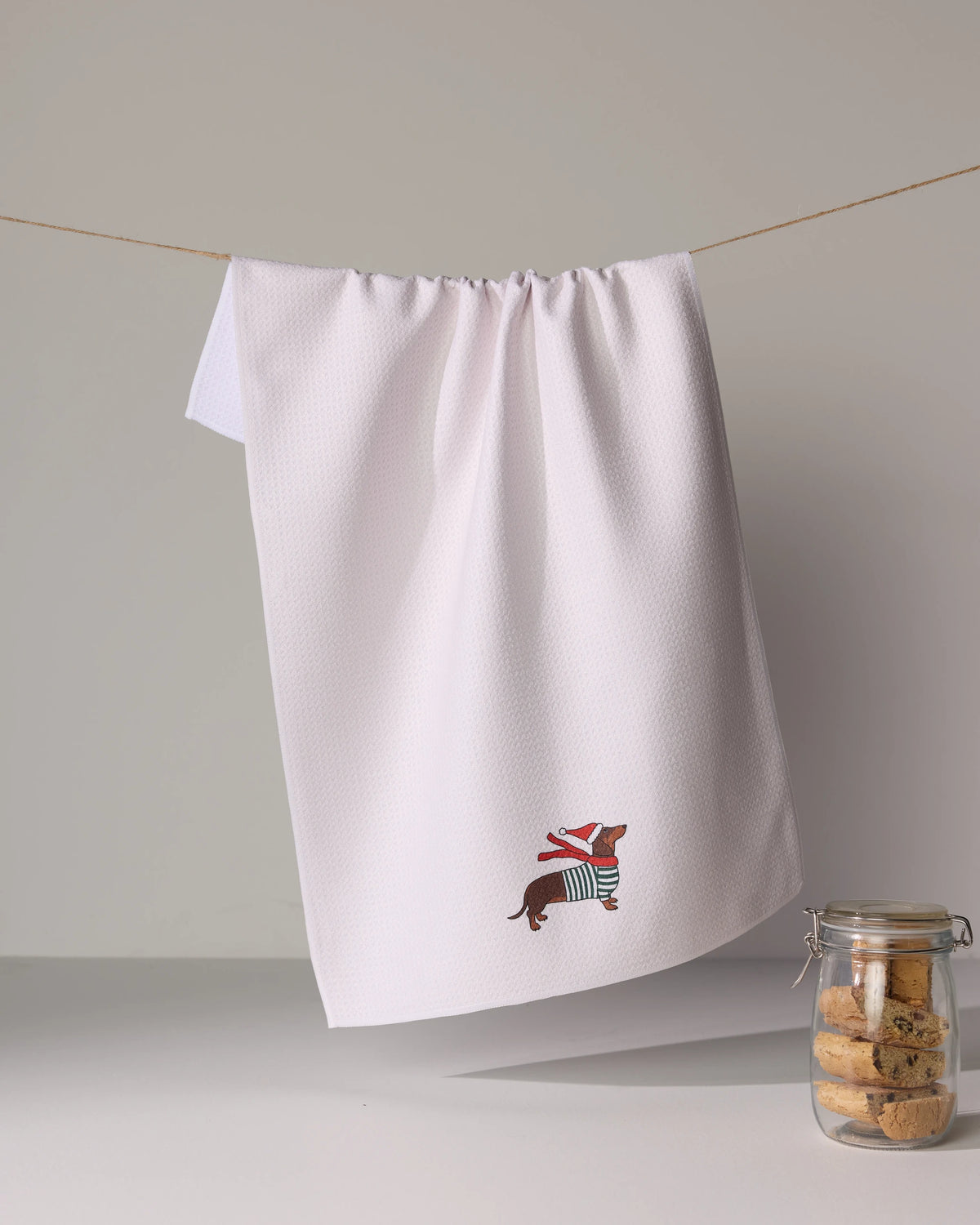 The Geometry Merry Dachshund is a quick-drying, recycled white tea towel with an embroidered dog in a striped sweater and scarf, shown hanging beside a cookie-filled glass jar.