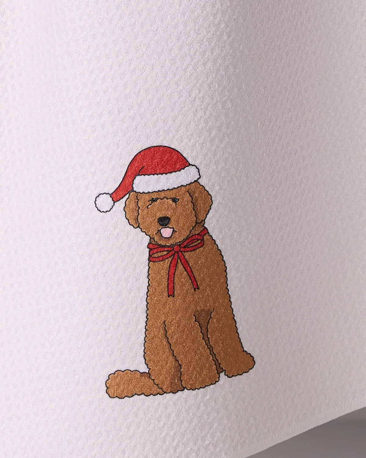 The Geometry Merry Doodle tea towel features a smiling brown dog in a red Santa hat and bow, printed on an ultra-absorbent towel made from recycled materials.