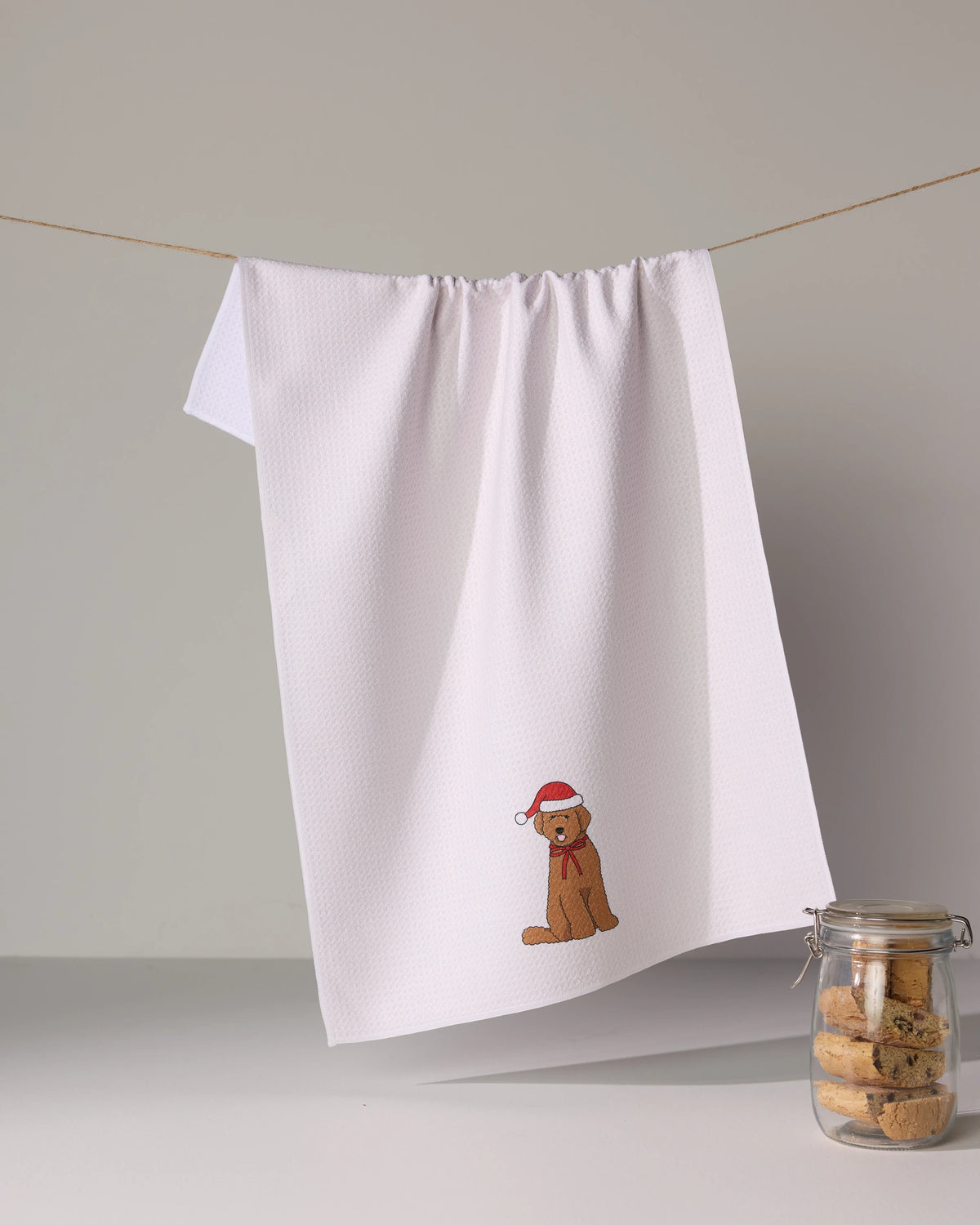 The Geometry Merry Doodle is a white tea towel featuring an embroidered dog in a Santa hat, made from ultra-absorbent recycled materials, hanging above a glass jar of cookies.