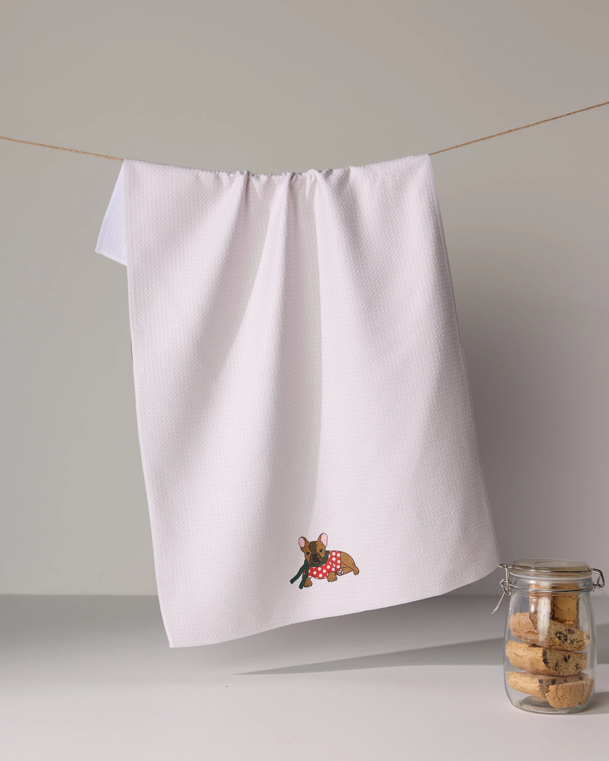 The Geometry Merry Frenchie ultra-absorbent white tea towel with an embroidered dog in a red polka dot bandana hangs on a string, with a jar of cookies on the right.