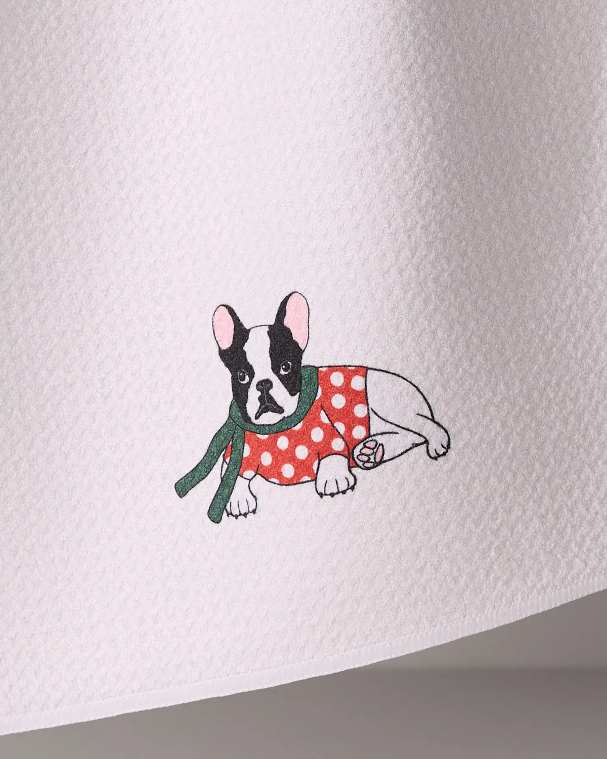 Geometry Merry Frenchie tea towel features a black and white French bulldog in a red polka dot sweater and green scarf, embroidered on ultra-absorbent textured white recycled fabric.