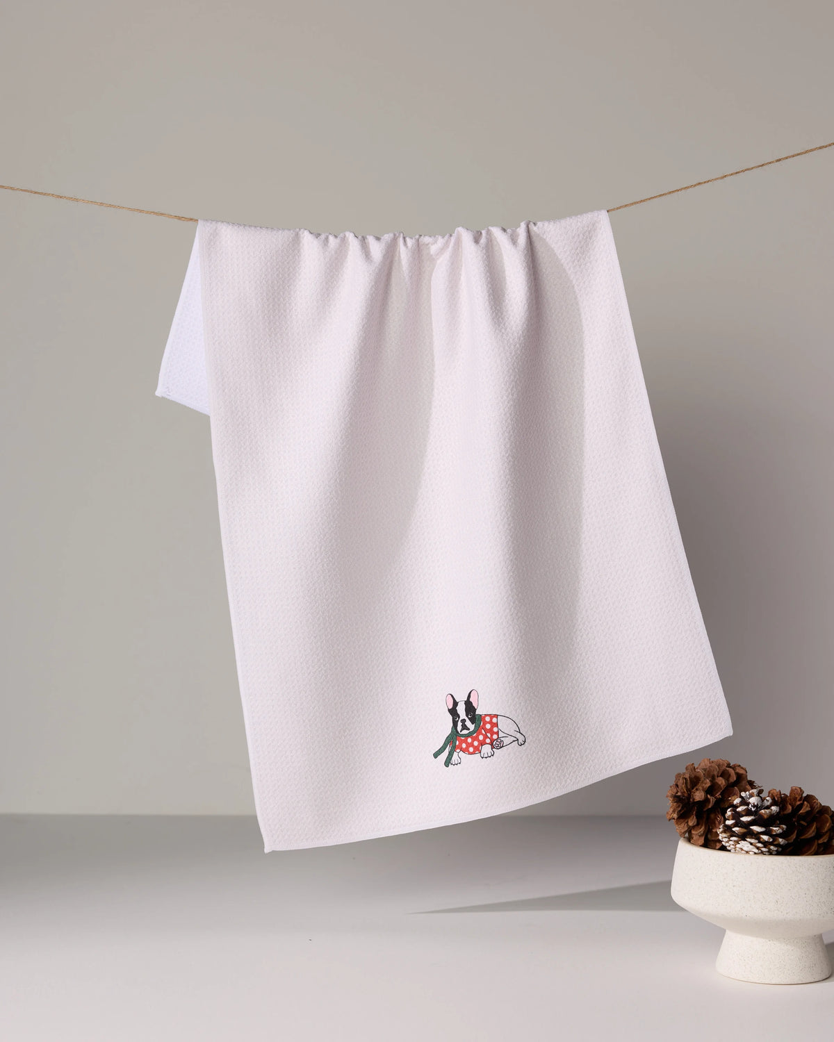 The Geometry Merry Frenchie, a white tea towel with a small embroidered bunny in a red polka-dot dress, hangs on a string beside a white bowl of pinecones on a neutral surface.