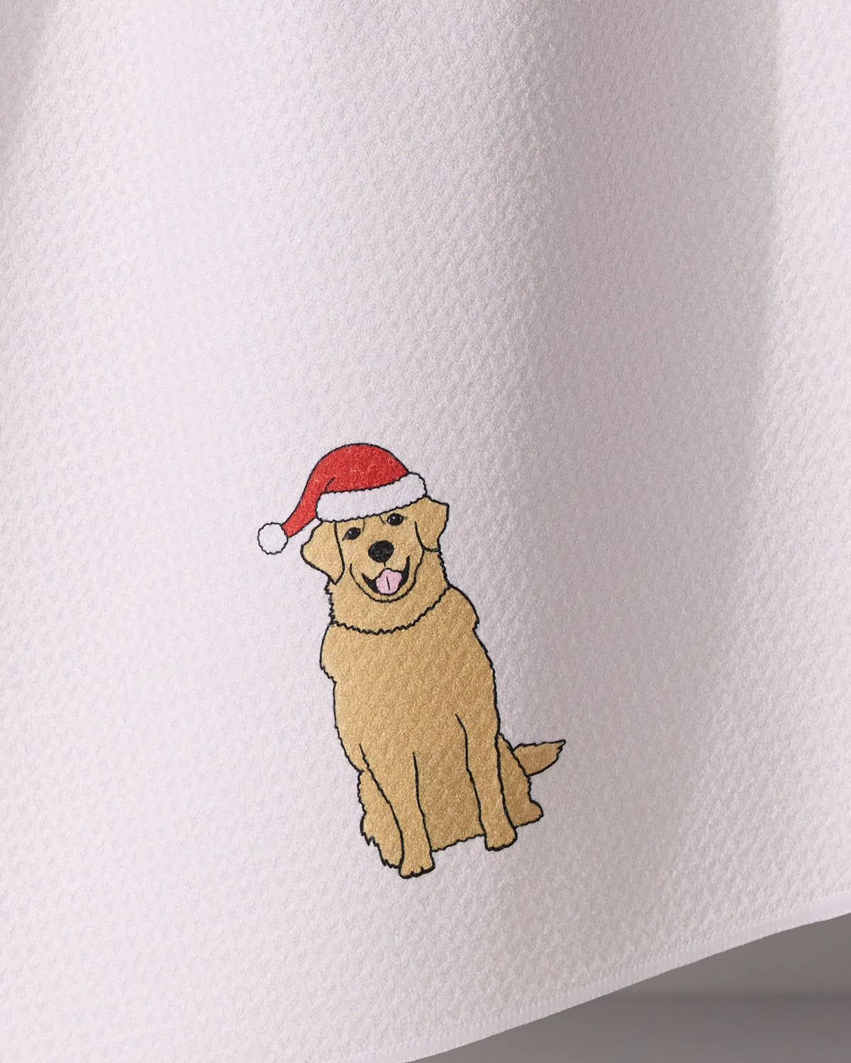 The Geometry Merry Golden is a white textured tea towel made from recycled materials, featuring a illustration of a smiling golden retriever in a red Santa hat.