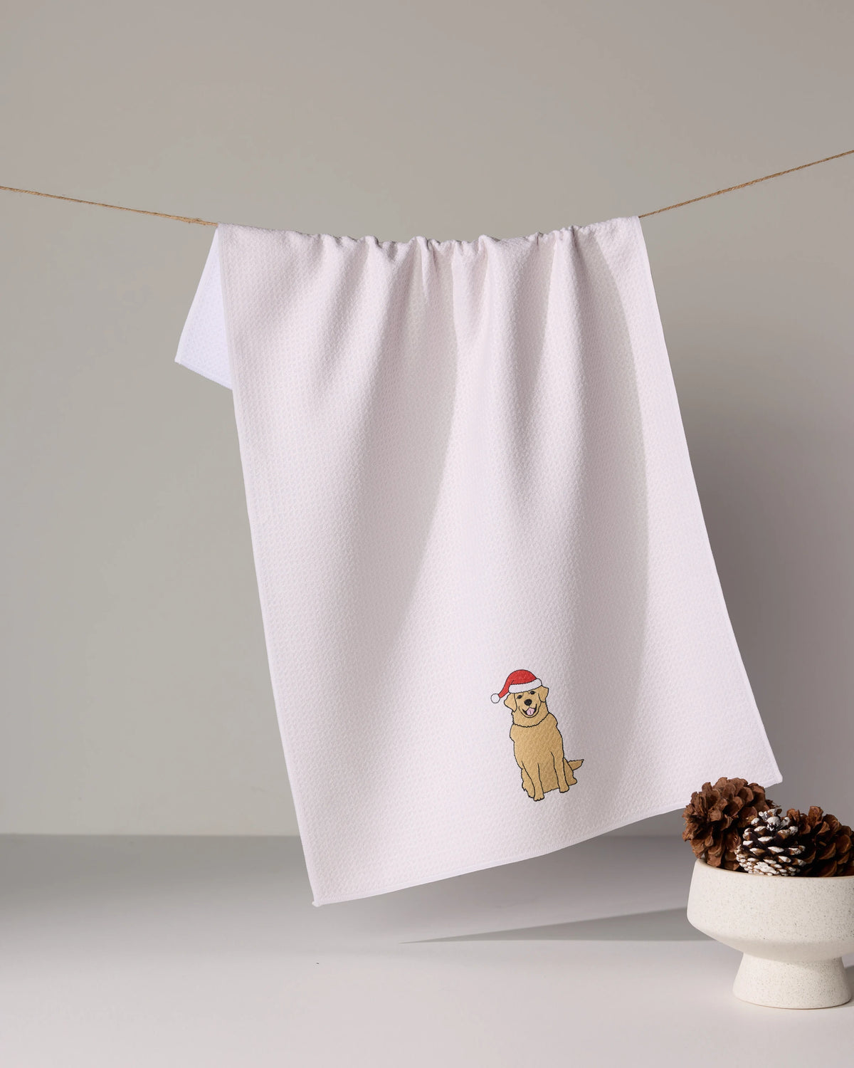 The Merry Golden tea towel by Geometry, made from recycled materials and embroidered with a dog in a red Santa hat, hangs on a string beside a small white bowl of pinecones.