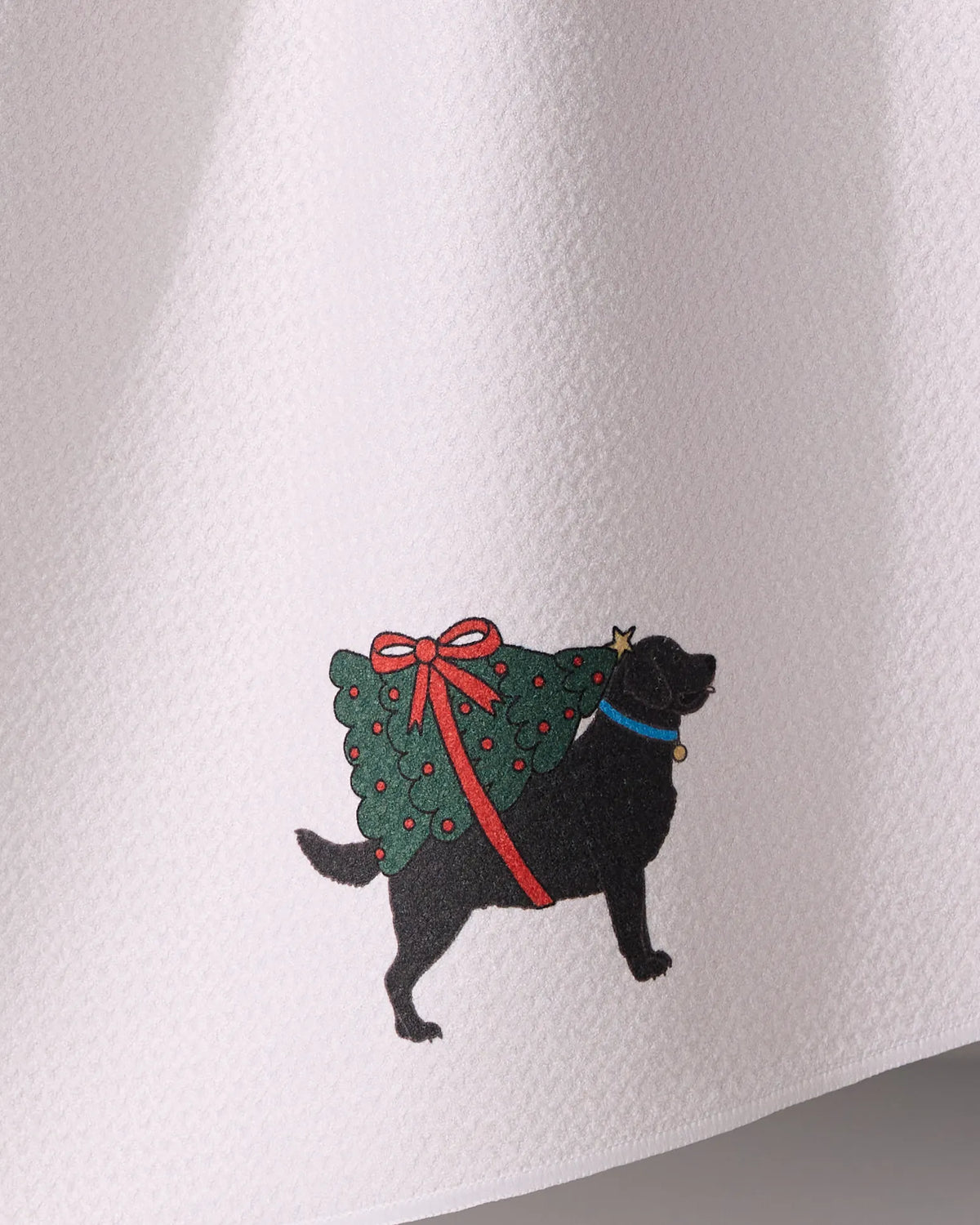 The Geometry Merry Labs kitchen towel features a black dog with a blue collar carrying a decorated Christmas tree, all set against a white textured background and made from ultra-absorbent recycled materials.