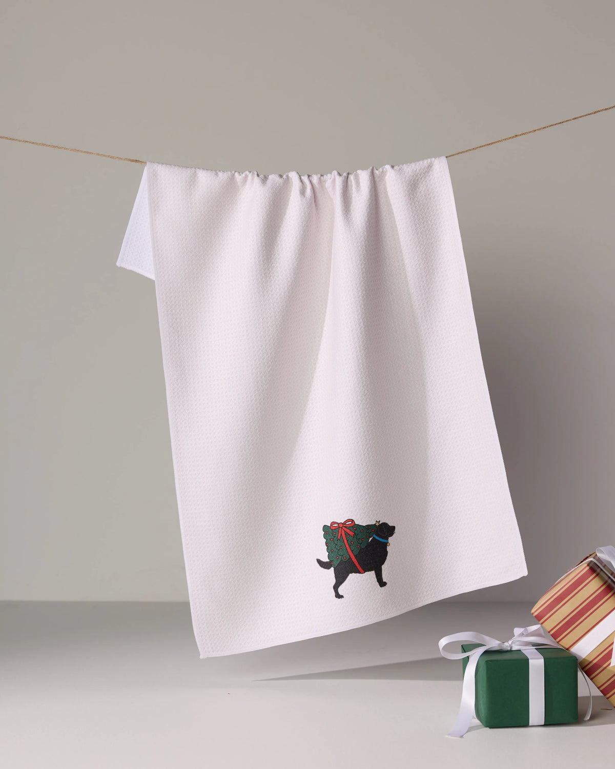 The Geometry “Merry Labs” white tea towel, featuring an embroidered black dog with a red bow, hangs on a clothesline with two wrapped gifts nearby on a white surface.