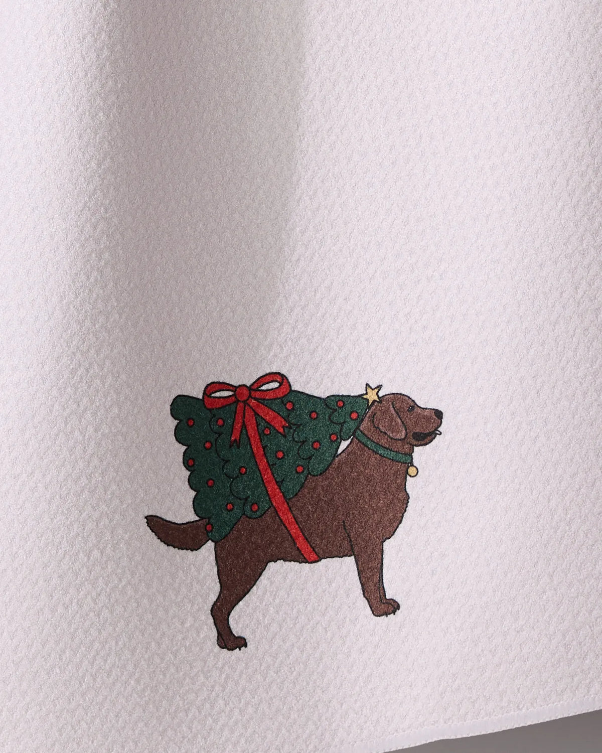 Geometry’s “Merry Labs” kitchen towel features a brown dog in a green Christmas tree costume with red decorations and a bow, printed on ultra-absorbent fabric.