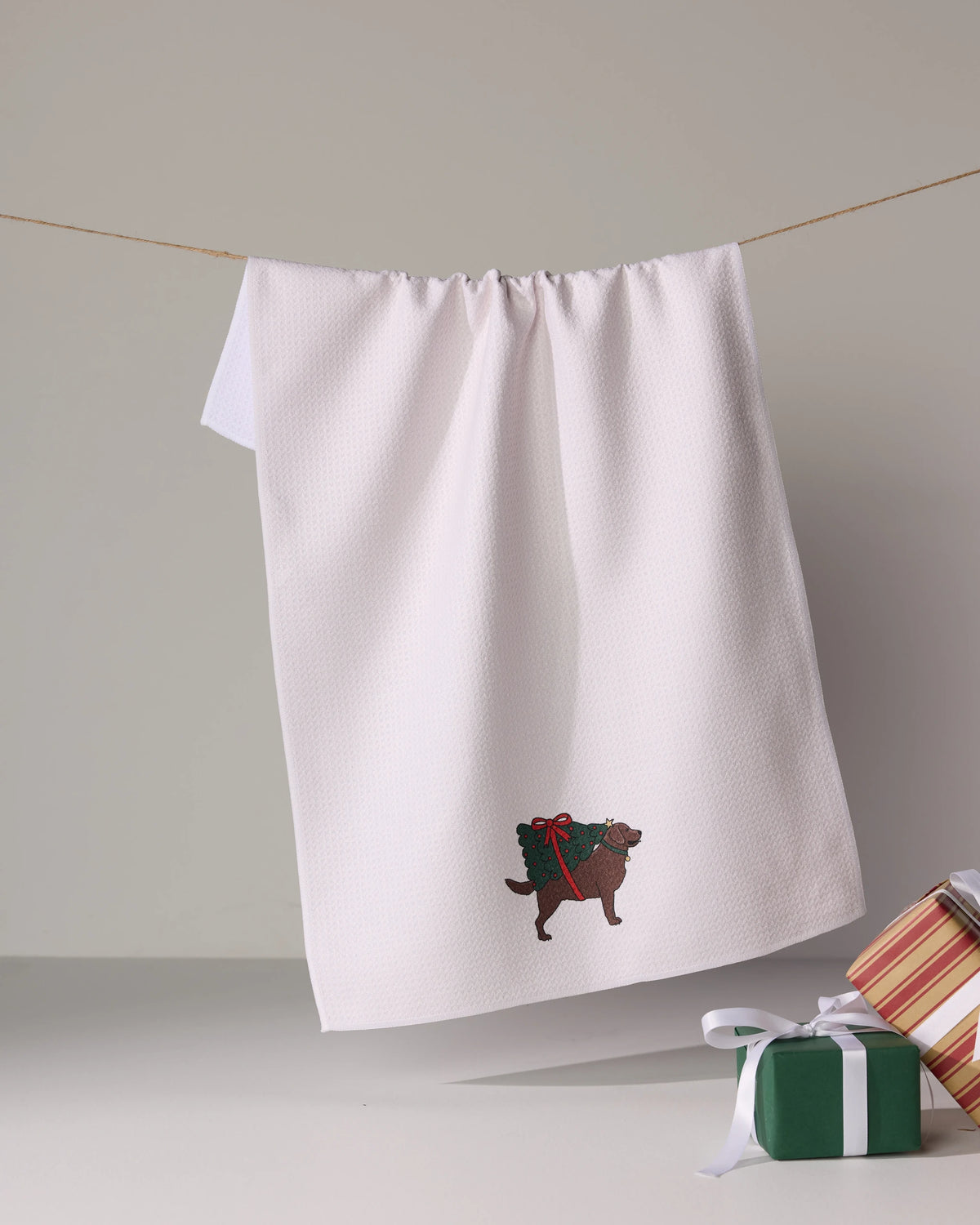 The Geometry Merry Labs tea towel features an embroidered dog with a green gift and red ribbon, plus two small presents, all on an ultra-absorbent white kitchen towel.