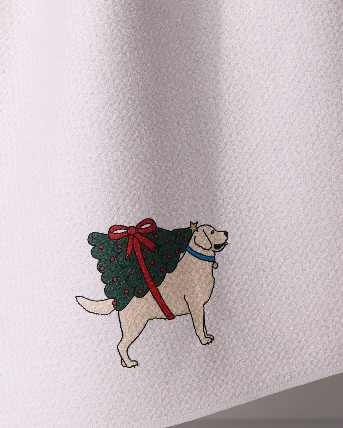 The Geometry Merry Labs kitchen towel features a cartoon dog carrying a Christmas tree decorated with red ornaments and a bow, printed on ultra-absorbent textured white recycled material.