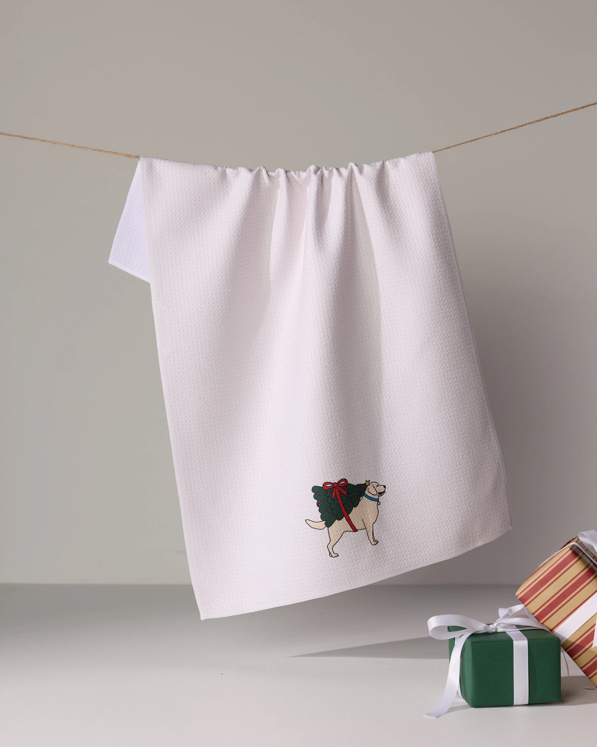 A Geometry Merry Labs white tea towel hangs on a string, embroidered with a dog carrying a holiday wreath. Gift boxes with ribbons sit in the bottom right corner.