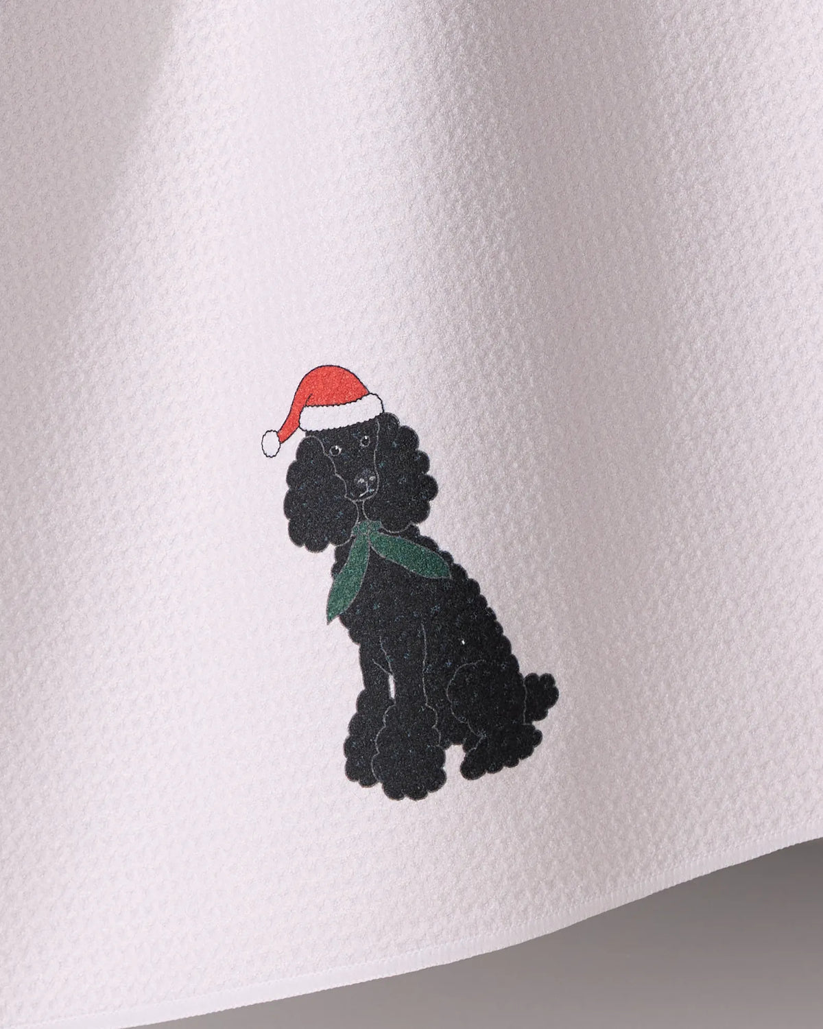 The Geometry Merry Poodle kitchen towel features a black poodle in a red Santa hat and green scarf, printed on an absorbent, recycled material with a textured white surface.