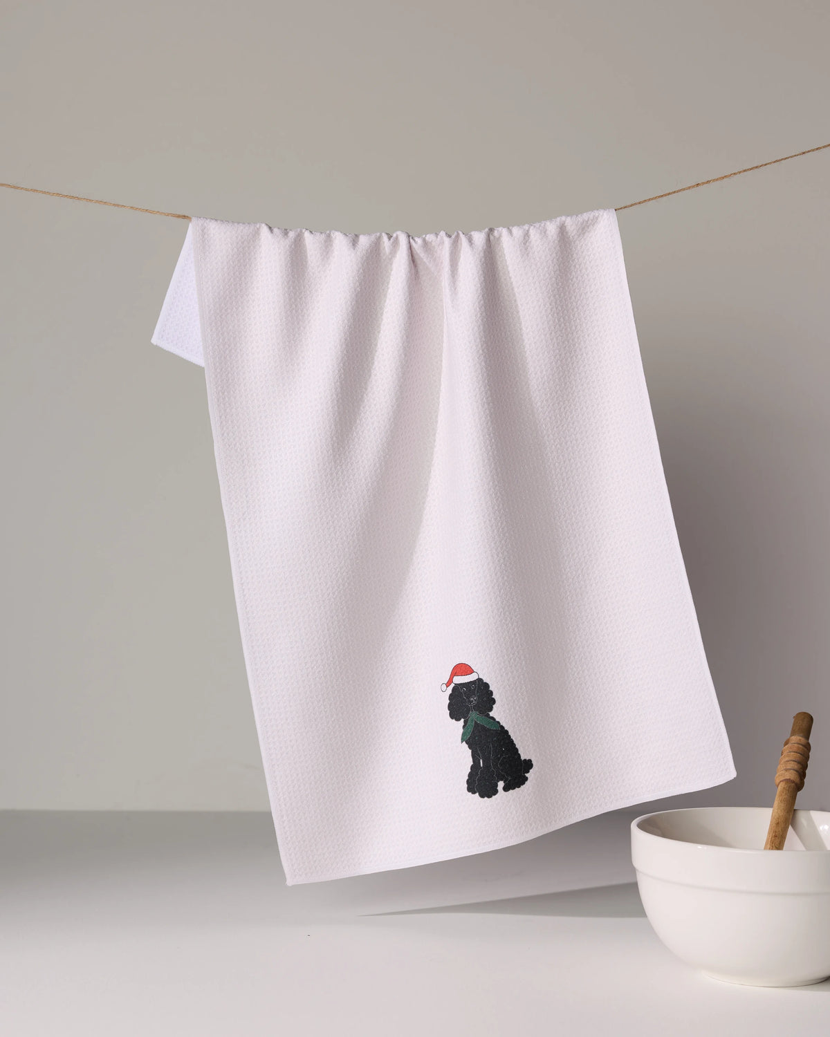 The Geometry “Merry Poodle” is a white absorbent kitchen towel featuring a small embroidered black dog with a red Santa hat, shown hanging above a white bowl with a honey dipper.