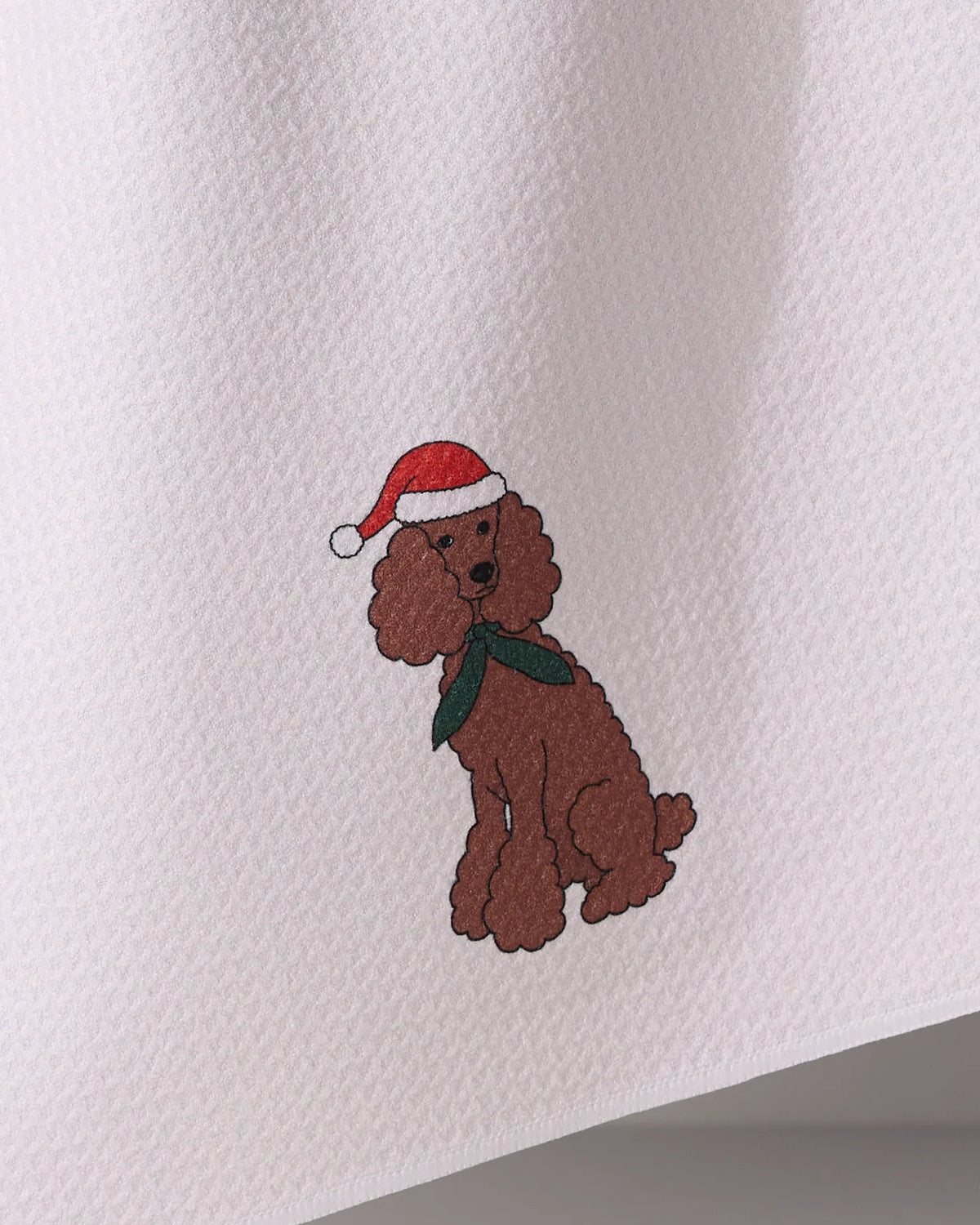 The Geometry Merry Poodle tea towel features an illustrated brown poodle in a red Santa hat and green scarf on textured white fabric made from recycled materials.