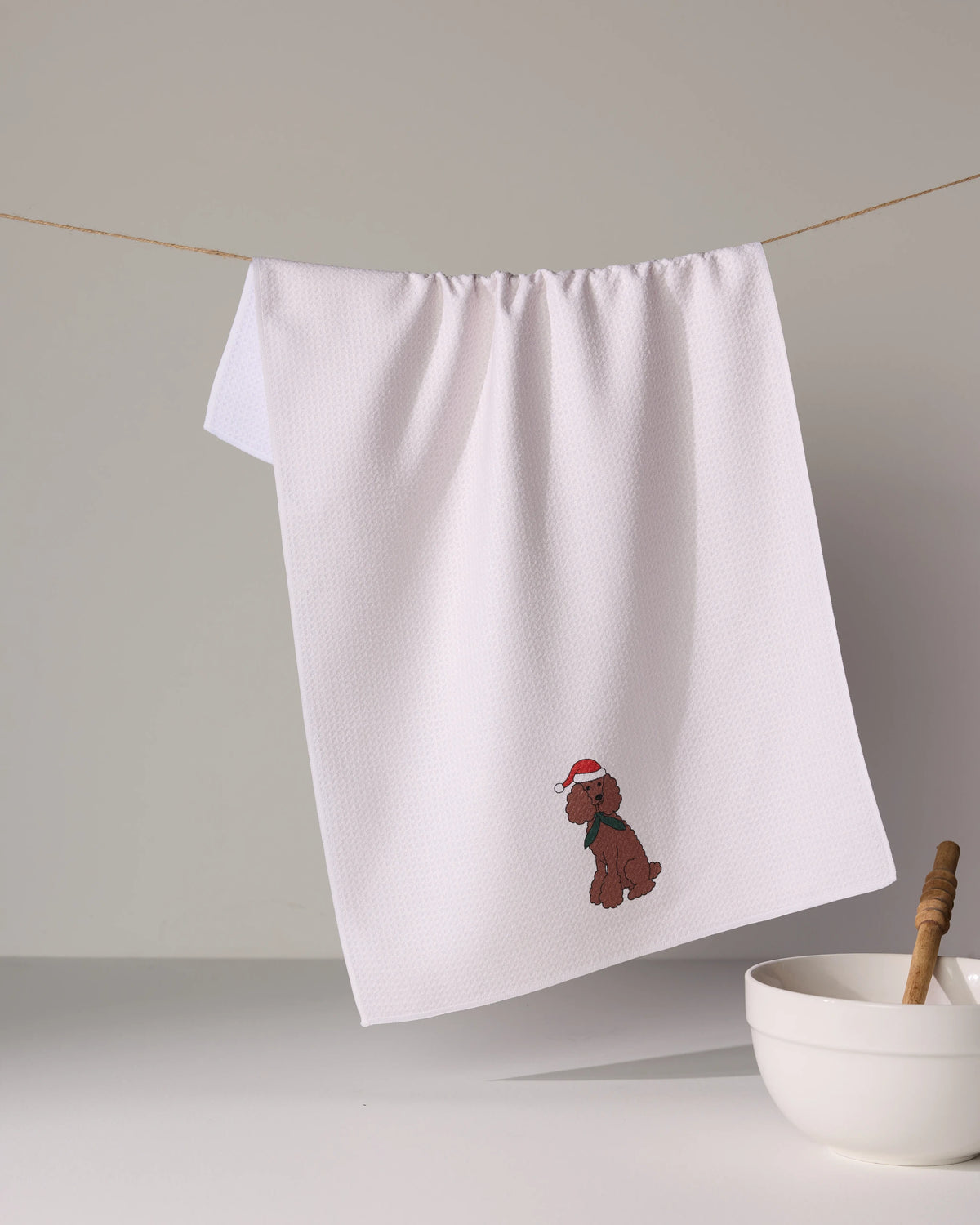 The Geometry Merry Poodle is a white tea towel featuring a small embroidered dog in a red Santa hat and green scarf. Made from absorbent recycled materials, it hangs above a bowl with a honey dipper.