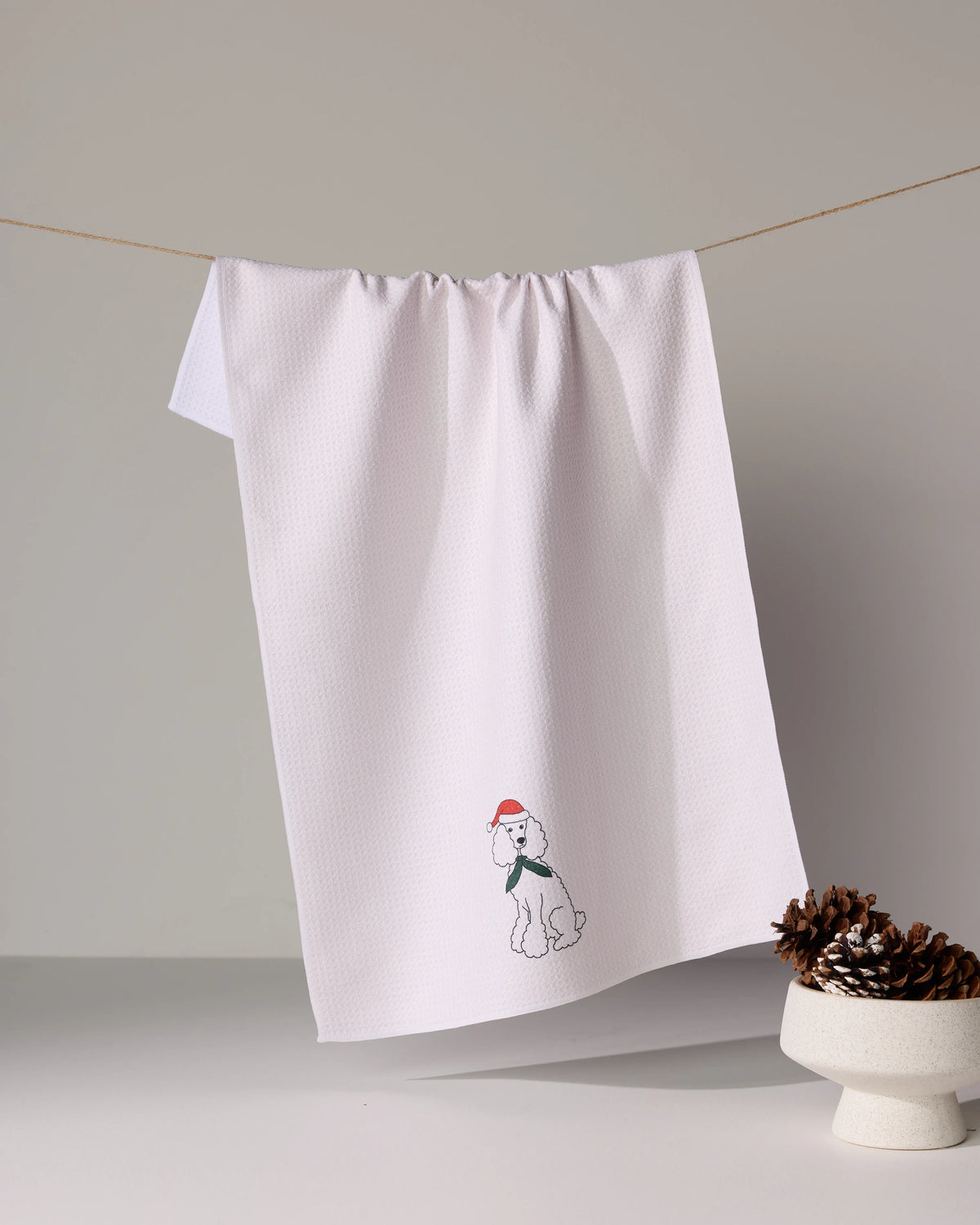The Geometry Merry Poodle white tea towel, featuring an embroidered dog in a red Santa hat, hangs on a line—an absorbent, festive addition to your holiday kitchen. Pine cones in a bowl complete the charming scene.