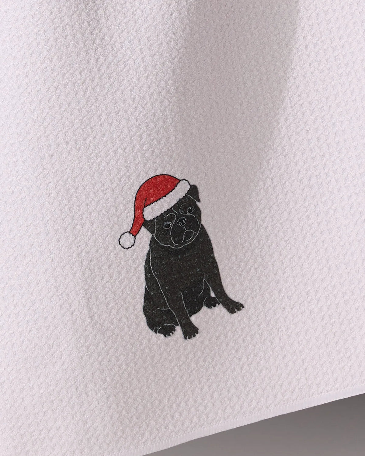 Geometry’s “Merry Pug” is a white textured tea towel made from recycled materials, featuring an embroidered black pug in a red and white Santa hat.