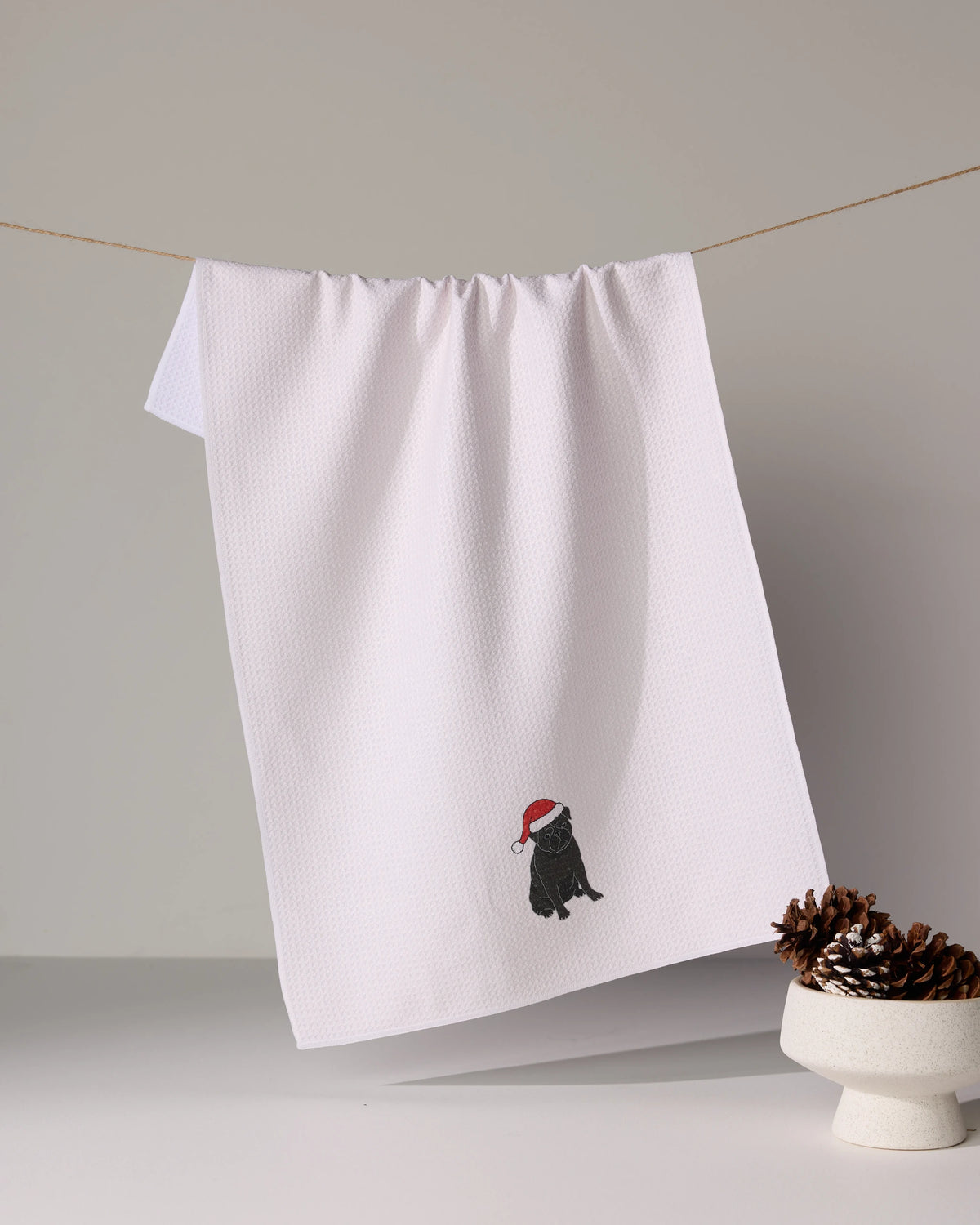 A white, ultra-absorbent Geometry kitchen towel featuring the “Merry Pug” design hangs on a line; beside it is a white bowl filled with pine cones.