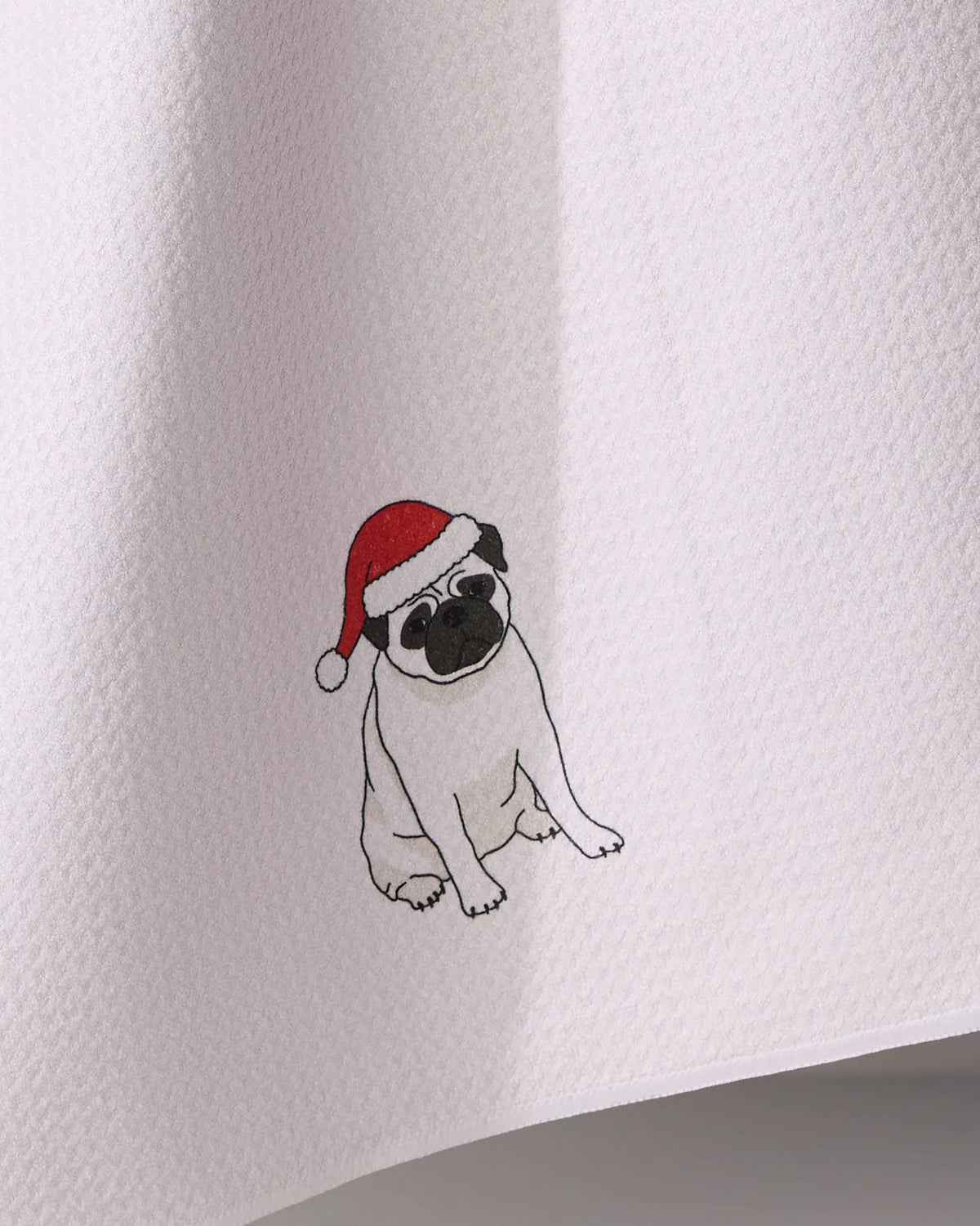 The Geometry Merry Pug tea towel features a white textured fabric with a pug in a red and white Santa hat illustrated near the bottom edge.