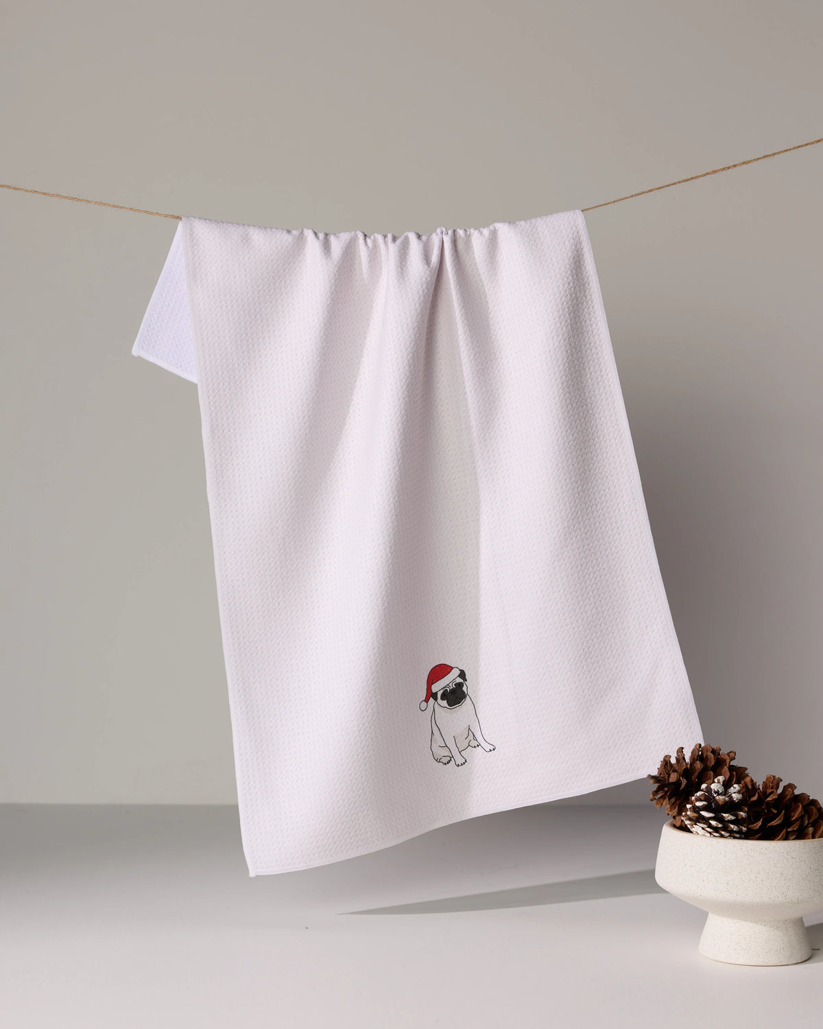 A white Geometry tea towel called “Merry Pug” hangs on a line, featuring a small embroidered pug in a red Santa hat. A bowl of pinecones sits in the foreground.