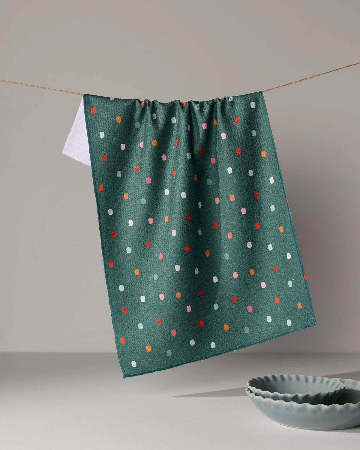 A Moss Tile Shuffle dark green tea towel with a pattern of small, multicolored rectangles hanging from a clothesline above two shallow bowls.