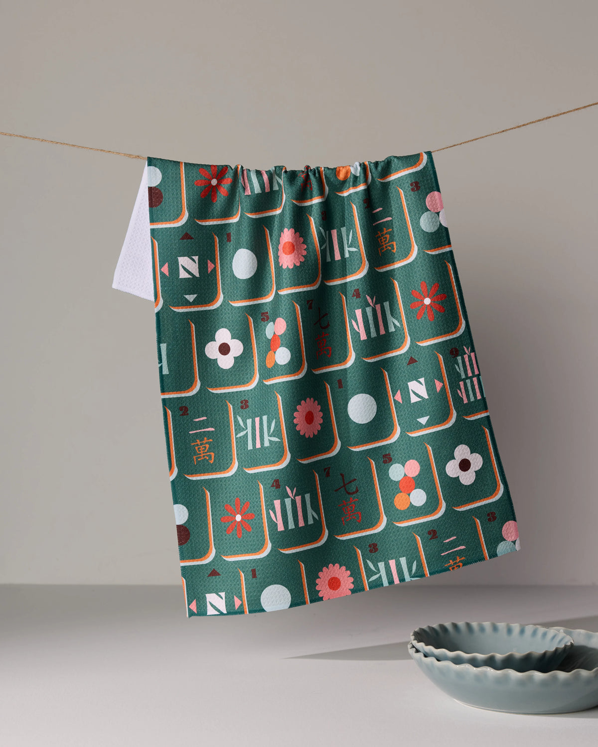 A quick-drying dark green Moss Tile Toss tea towel patterned with various mahjong tiles and floral icons hanging on a string.