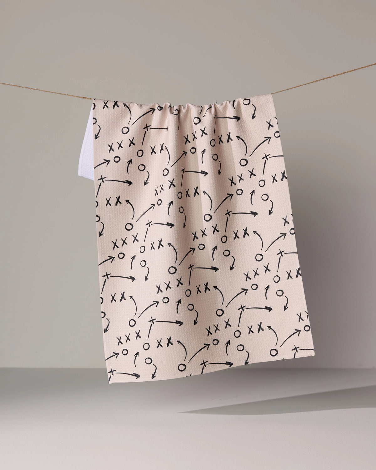 Pale pink Geometry Playbook tea towel patterned with black sports play diagrams hanging on a clothesline.
