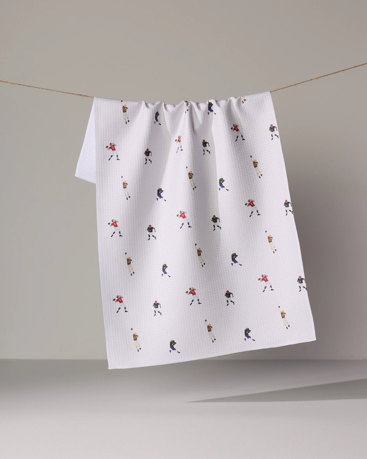 A quick-drying white Roster tea towel hanging on a string, patterned with small illustrations of football players in various uniforms.