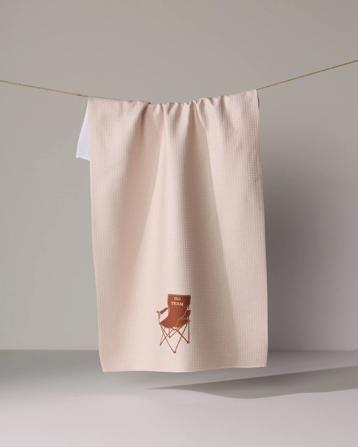 Geometry Tailgate tea towel in pale pink waffle-weave hanging on a line, featuring an embroidered brown graphic of a folding chair with the text GO TEAM.