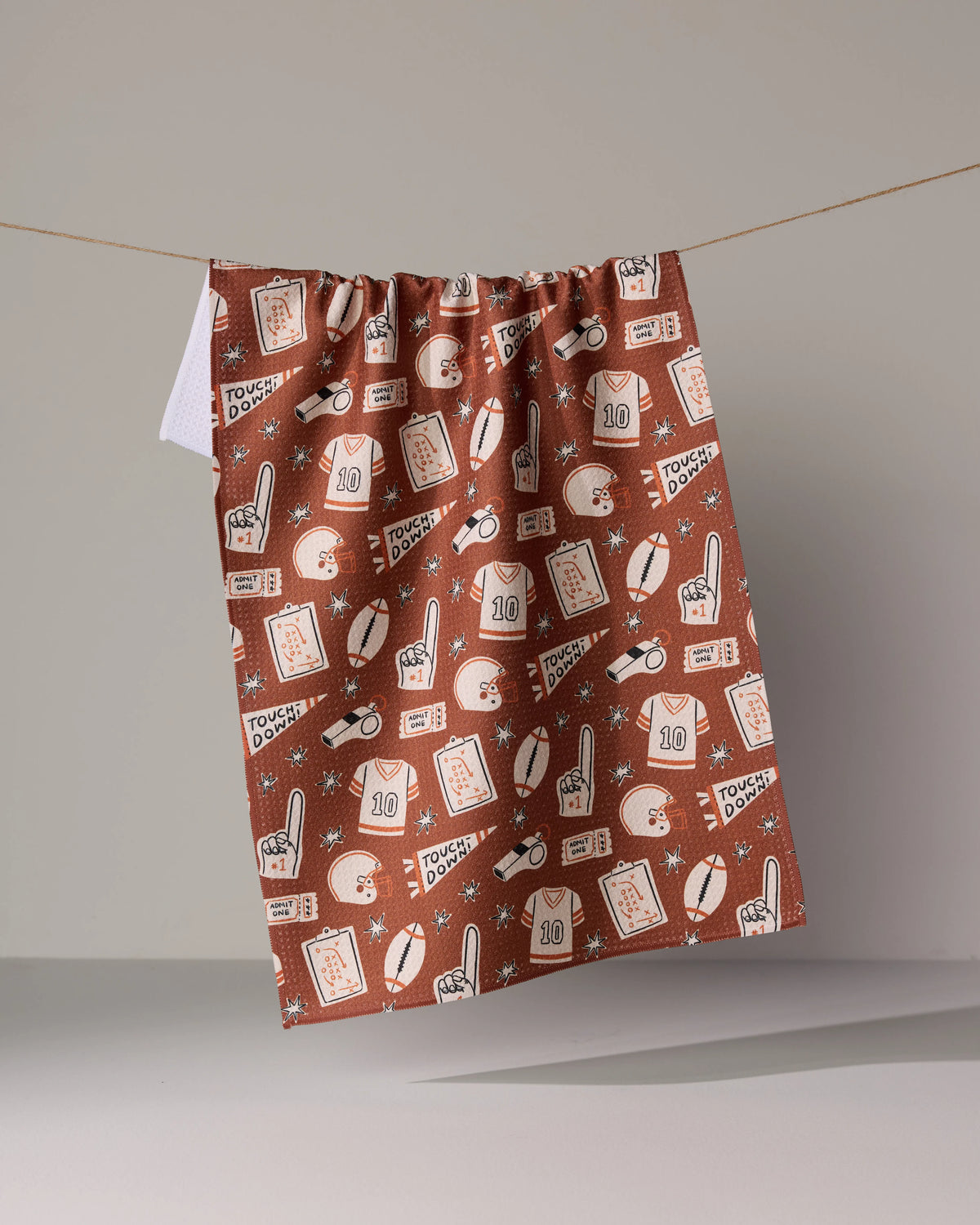 The Team Huddle brown tea towel features a repeating pattern of football-themed icons, including jerseys, helmets, footballs, whistles, tickets, and clipboards.