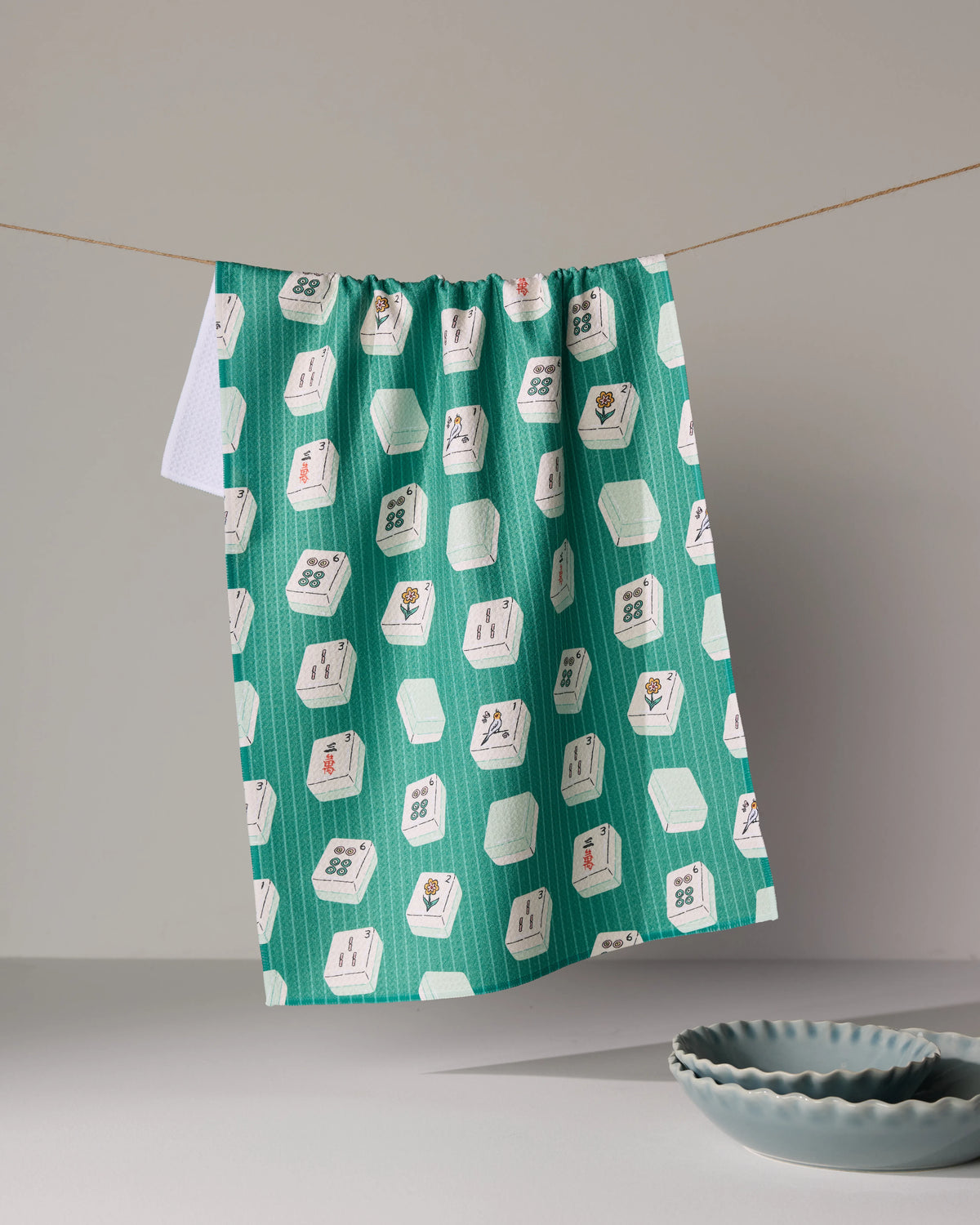 A Geometry Tile Flow kitchen towel in teal with a repeating mahjong tile pattern hangs on a clothesline above two ceramic bowls.