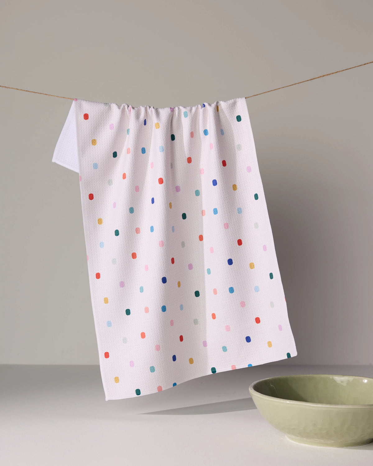 Geometry Tile Shuffle white waffle-weave kitchen towel featuring a pattern of colorful, pill-shaped dots hanging from a string above a green bowl.