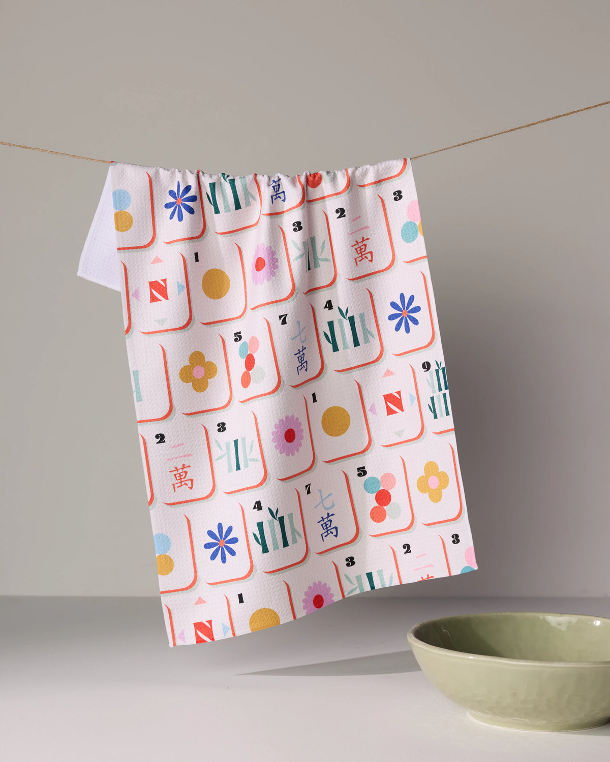 Geometry Tile Toss waffle-weave kitchen towel featuring a colorful pattern of mahjong tiles, hanging on a string above a green bowl.