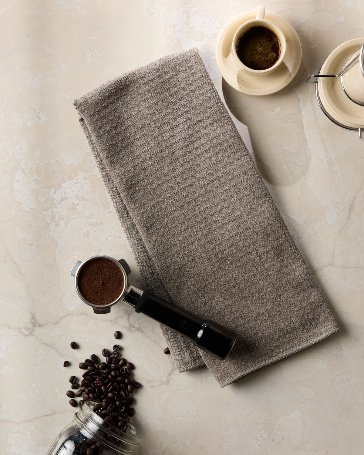 A folded ultra-soft waffle weave Geometry Pebble Hand Towel on a marble surface, with a cup of espresso, scattered coffee beans, and a portafilter nearby.