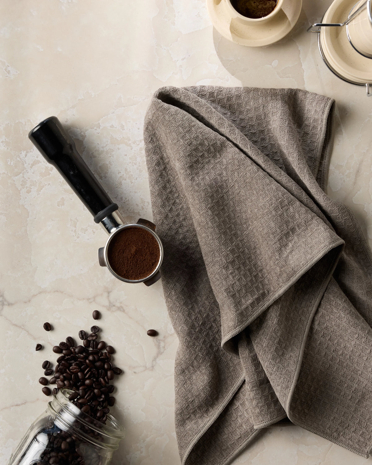 Coffee beans spill from a jar beside a tamper filled with ground coffee and the ultra-soft Geometry Pebble hand towel, crafted from recycled materials, on a marble countertop.