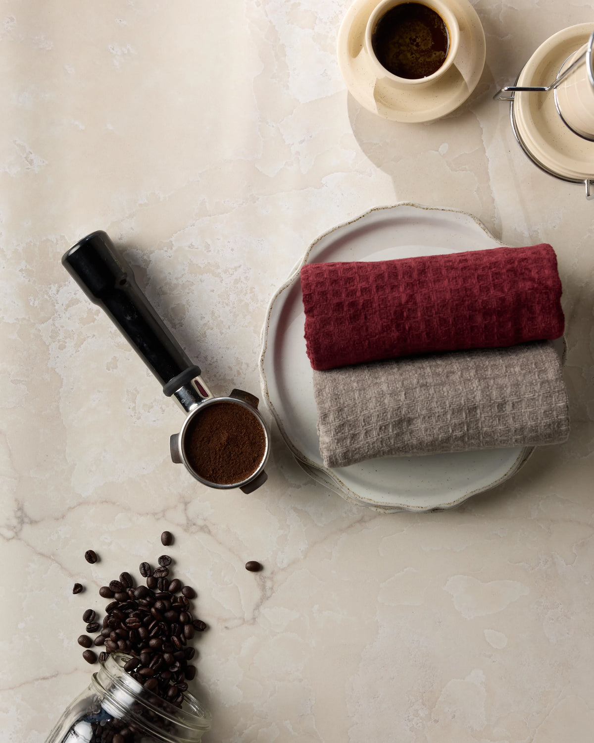 A portafilter with ground coffee, two Geometry Pebble ultra-soft waffle weave hand towels on a plate, a cup of espresso, and scattered coffee beans on a light marble surface.