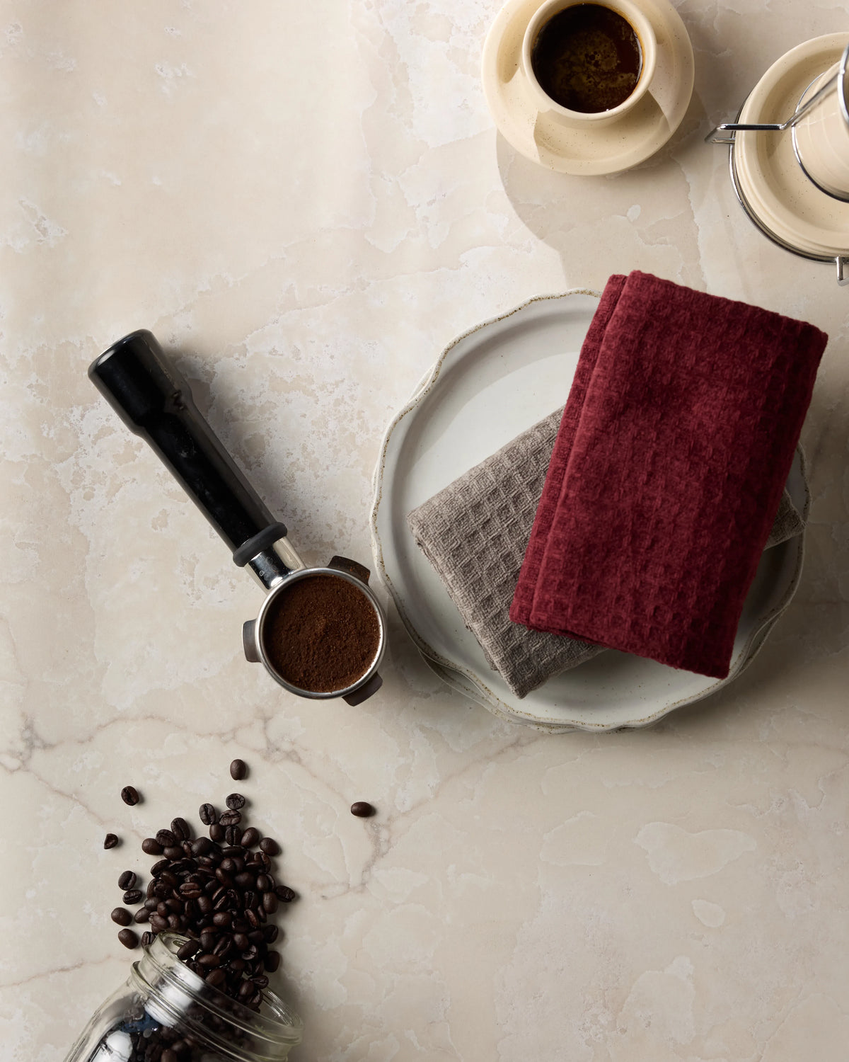 Coffee beans, a portafilter, an espresso cup, and the Geometry Merlot hand towel folded on a plate are artfully arranged on a light marble surface.