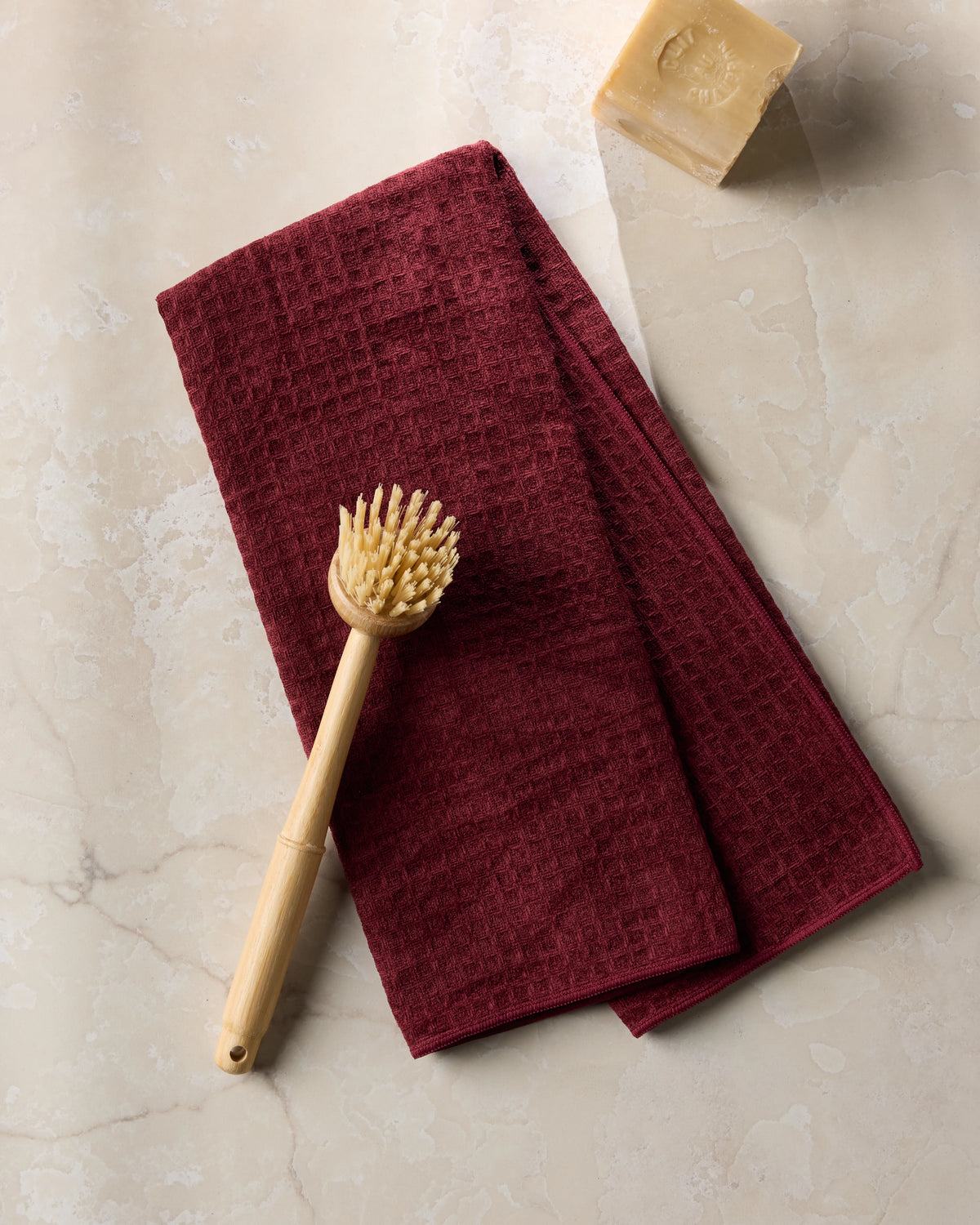 A wooden dish brush rests on a folded Geometry Merlot hand towel, with a bar of soap nearby on a light marble surface.