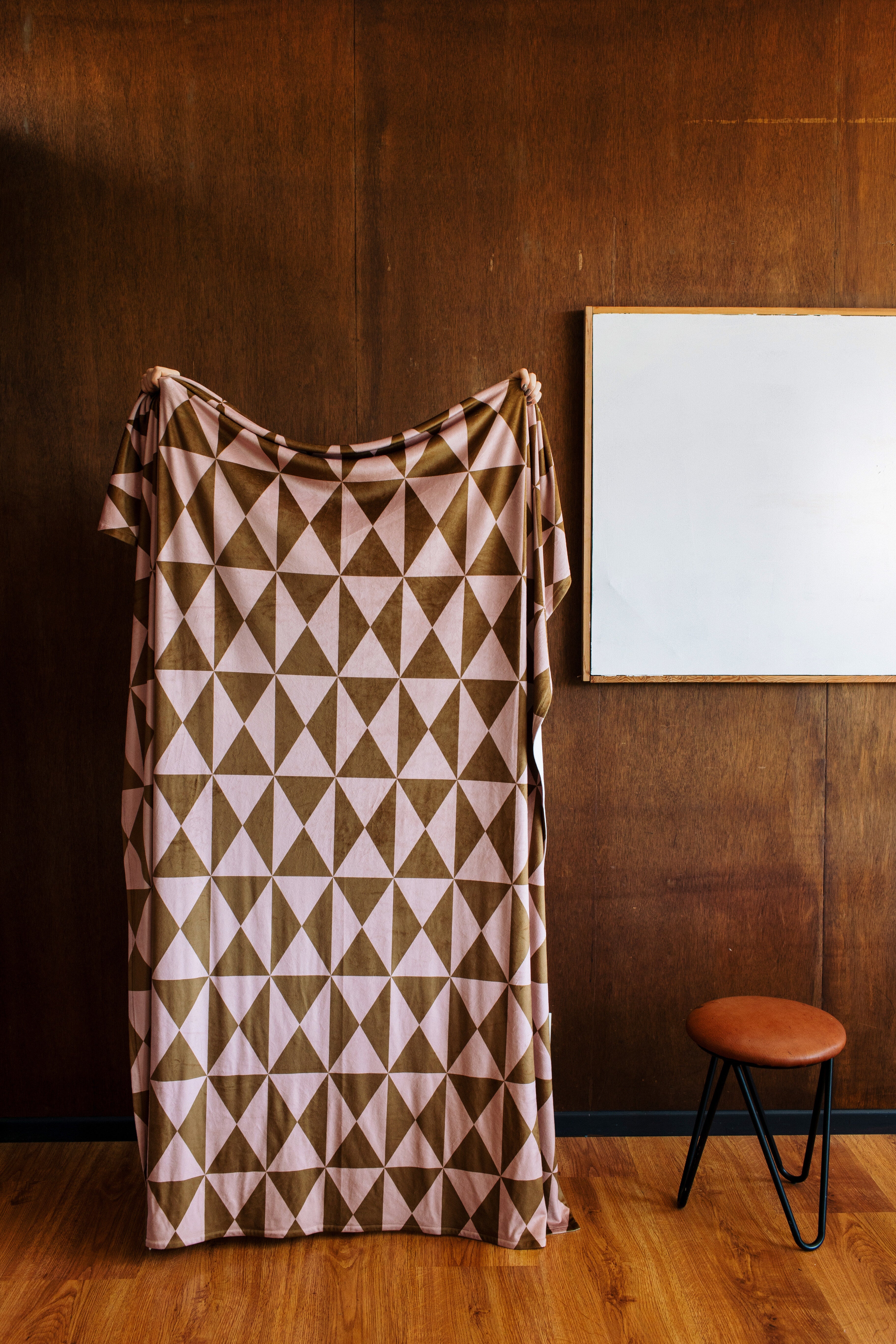 The Geometry "Bronze" Butter Blanket, featuring brown and pink triangles, hangs against a wooden wall beside a blank white canvas and a small round stool with a leather seat and black metal legs on a wooden floor.