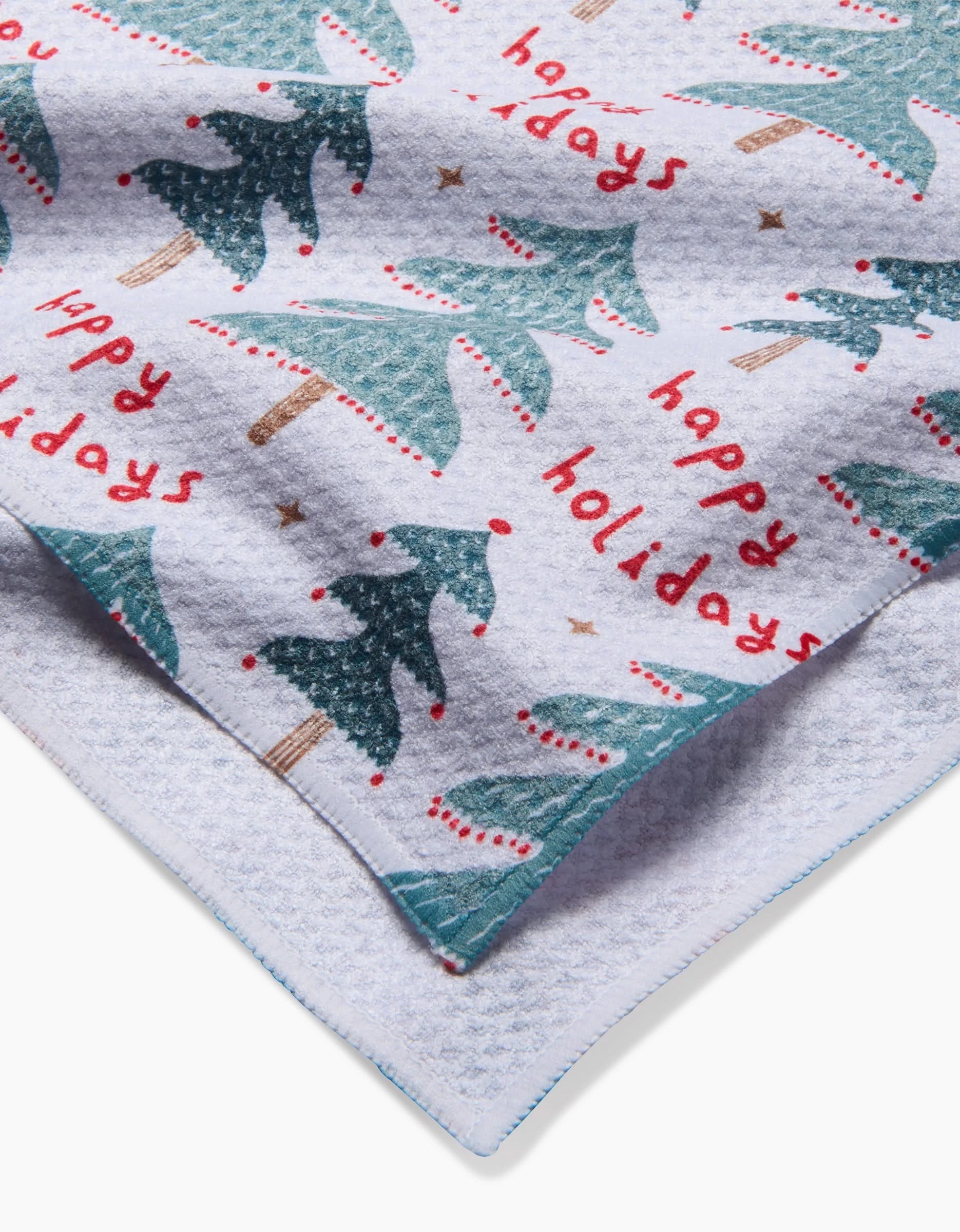 A close-up of the Roxy Marj Berry Happy Holidays Luxe Hand Towel, featuring green Christmas trees, red dots, happy holidays text, quick-drying Geoweave technology, and blue-stitched edges.
