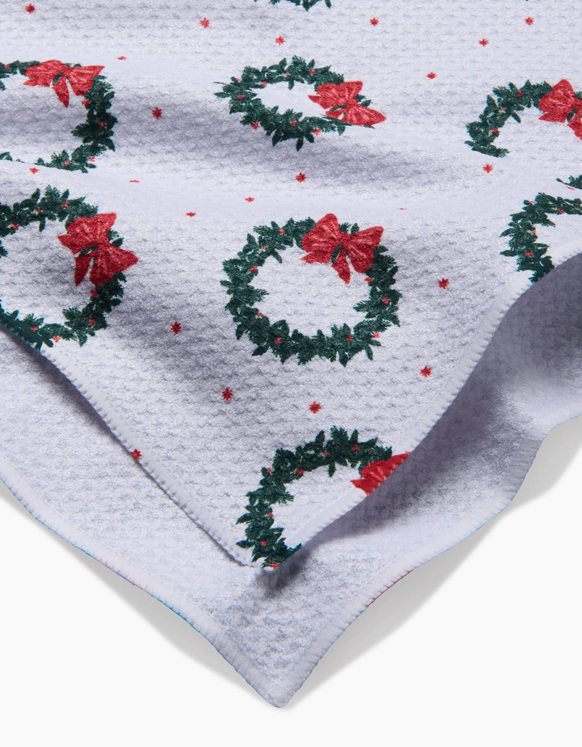 The Ash Cascade Bow Wreath Luxe Hand Towel features a white textured fabric with green Christmas wreaths, red bows, and small red stars. Quick-drying material is shown folded to highlight its corner and layered design.