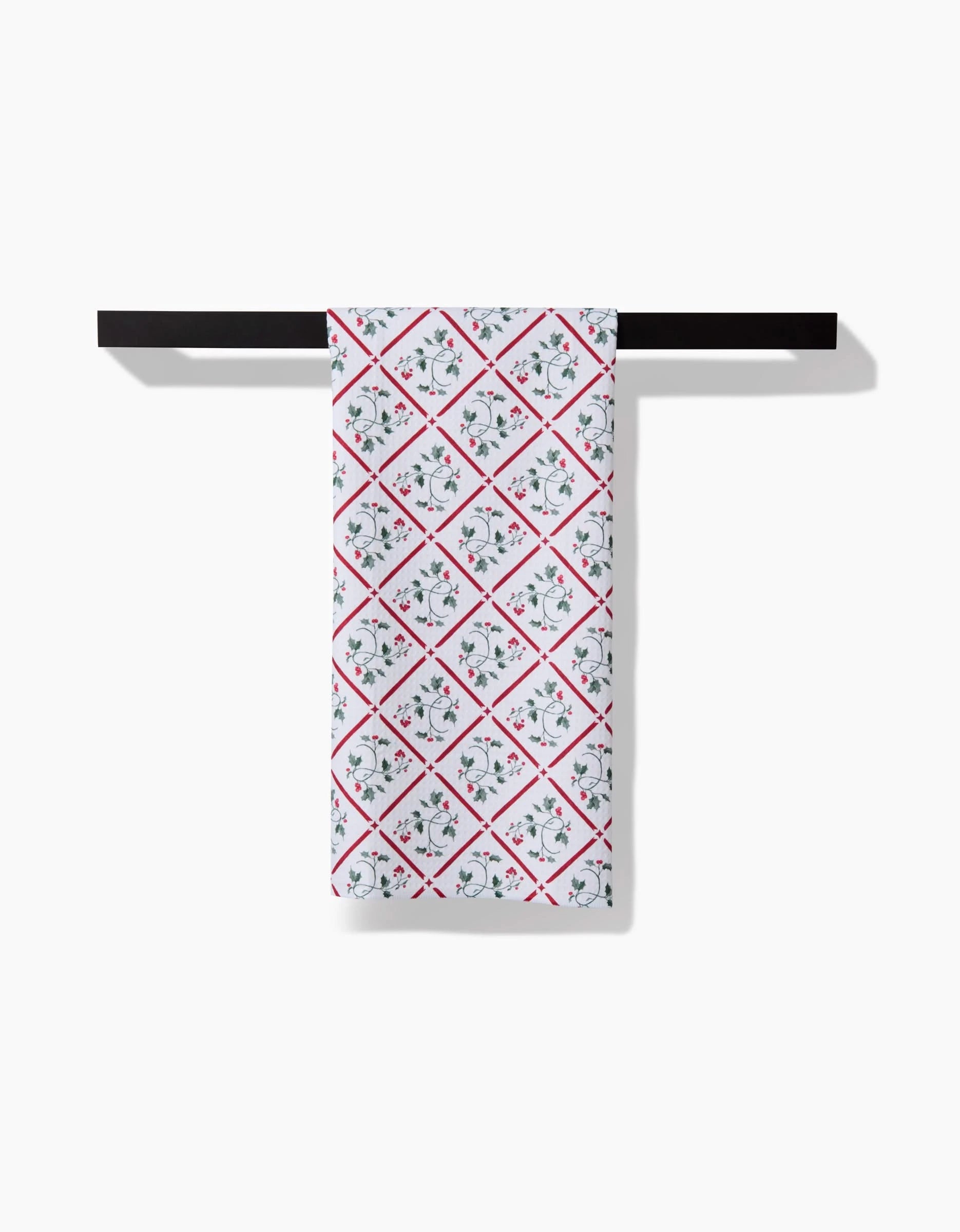 The Aubrey Fairchild Christmas Mistletoe Trellis Luxe Hand Towel, showcasing red diamond and green floral designs on white, hangs folded on a black towel bar. Its Geoweave technology ensures quick drying and lasting freshness.