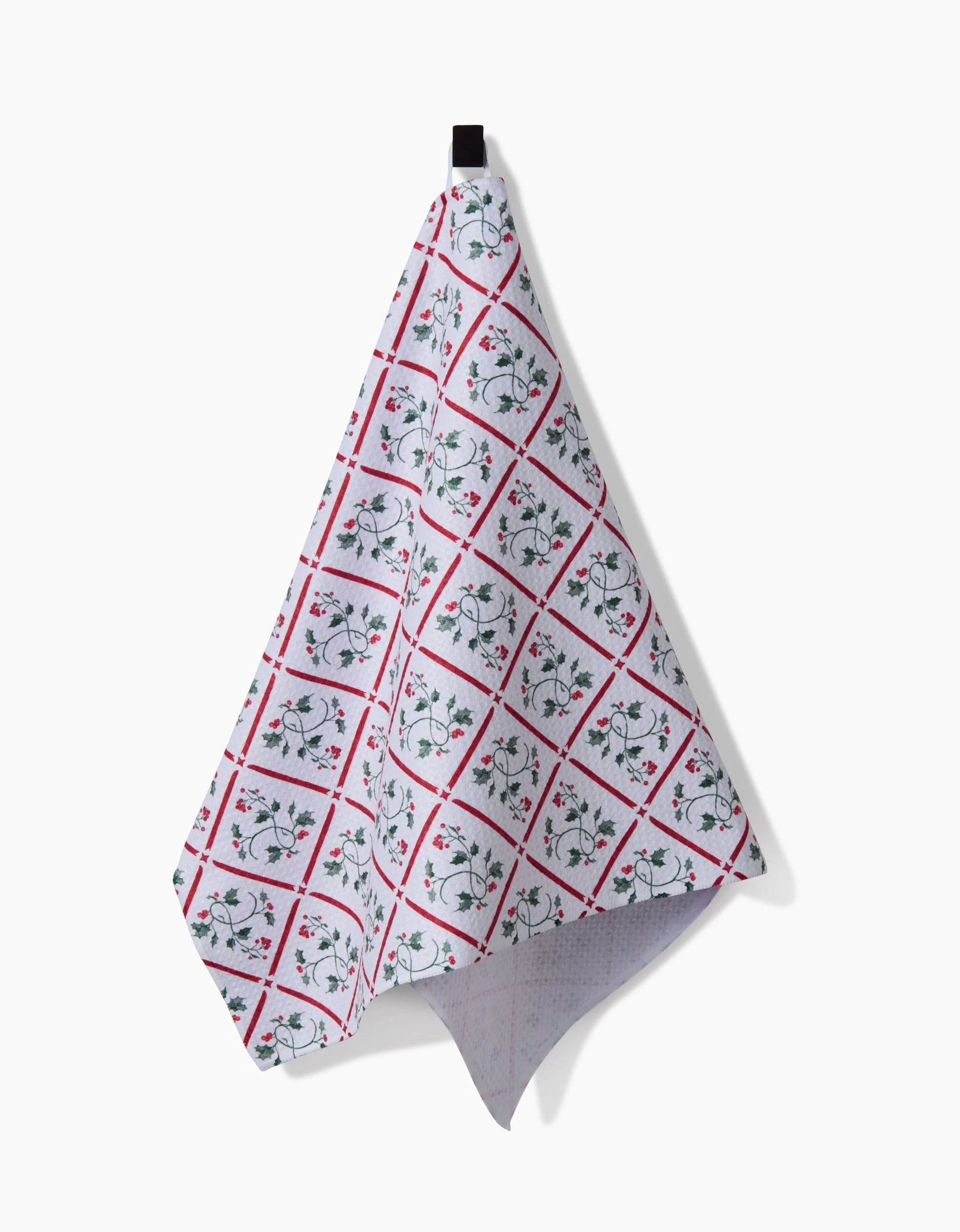 The Aubrey Fairchild Christmas Mistletoe Trellis Luxe Hand Towel, with a red diamond grid and green floral design in quick-drying Geoweave, hangs from a black loop against a plain white background.