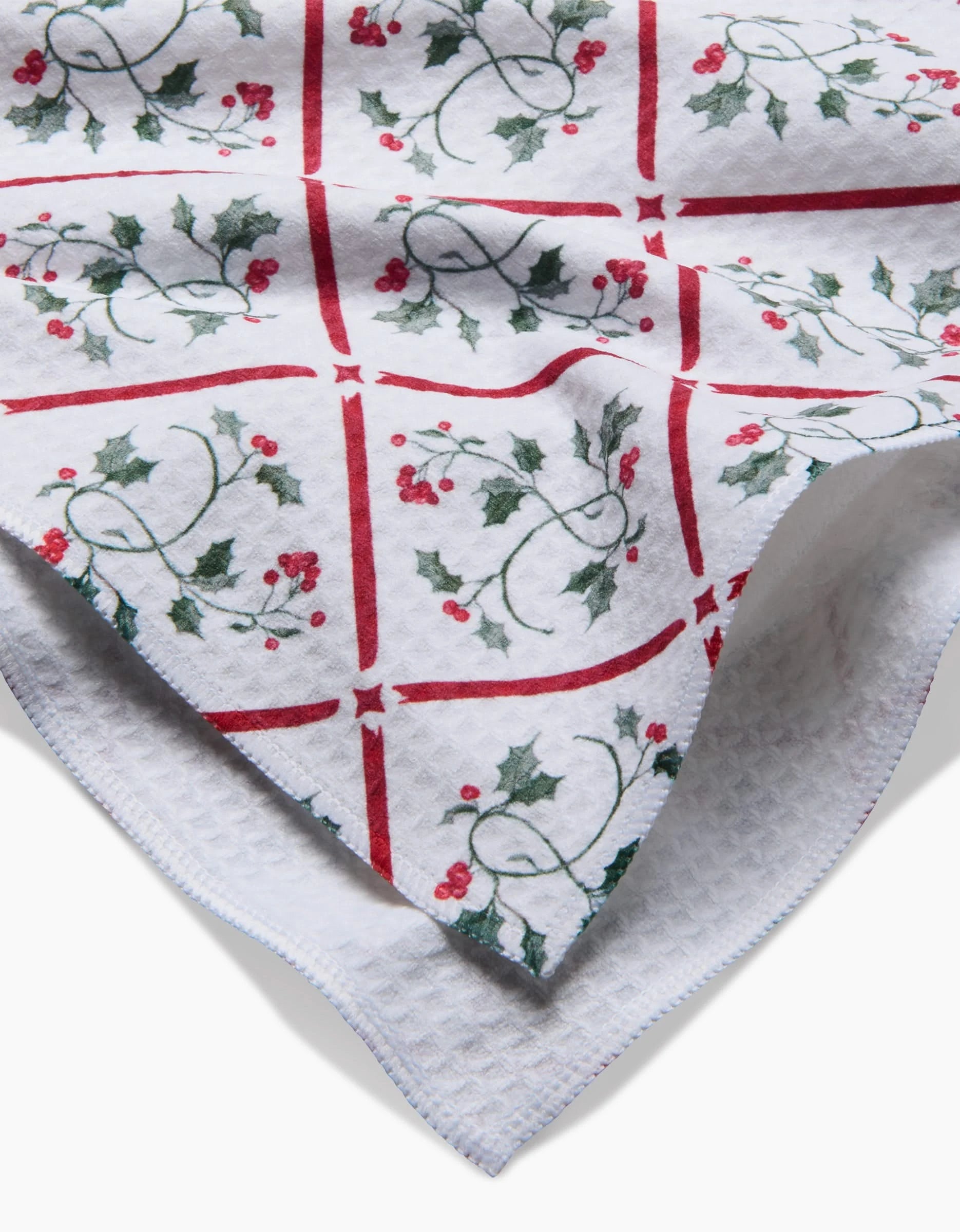 A close-up of the Aubrey Fairchild Christmas Mistletoe Trellis Luxe Hand Towel, showcasing red square lines with green holly and berries. Made with quick-drying Geoweave technology; towel’s corner is folded over.