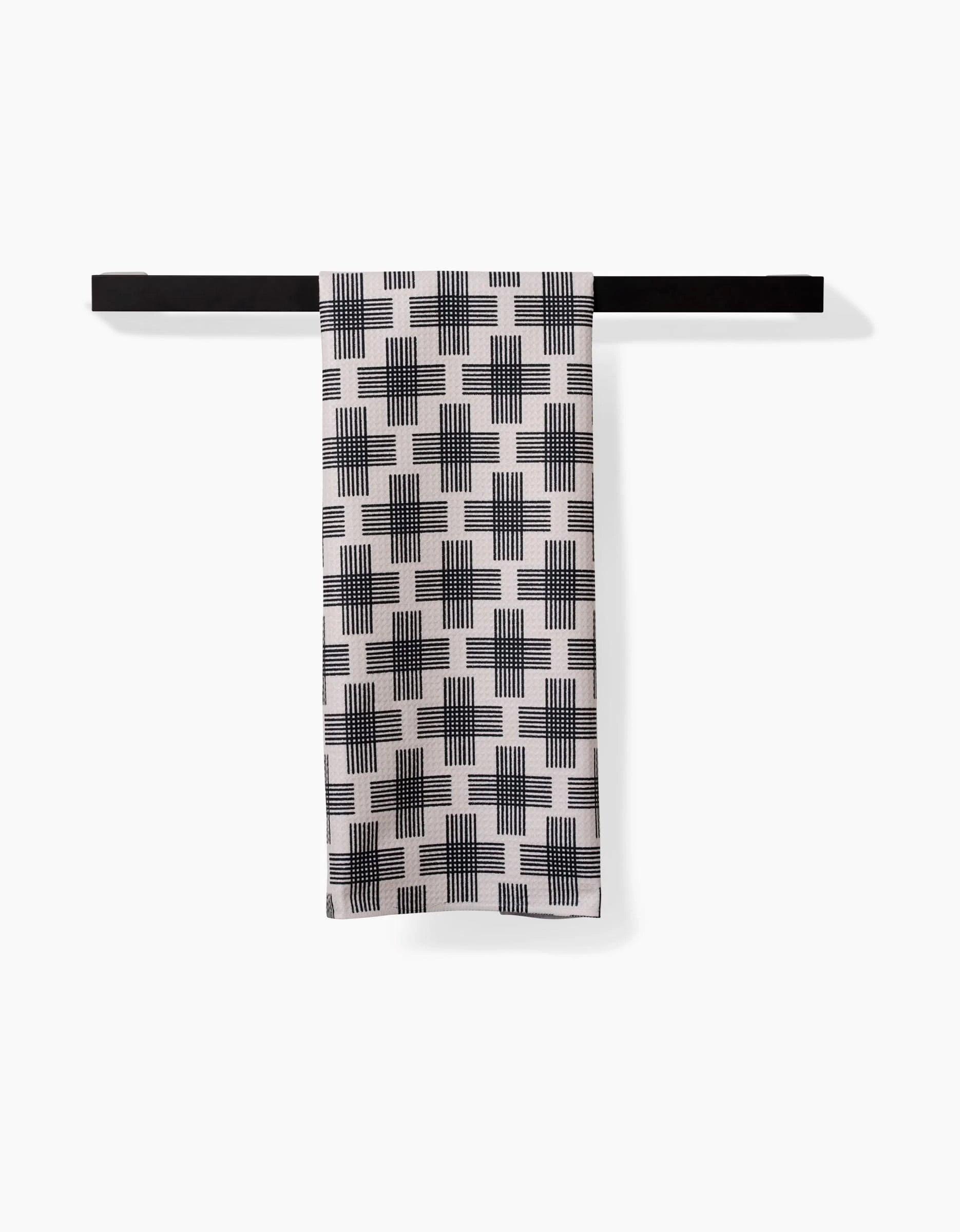The Rosa Kichline Cross Hatch Luxe Hand Towel, featuring a black geometric grid design and ultra-absorbent Geoweave technology, hangs neatly from a sleek black towel bar against a plain white background.