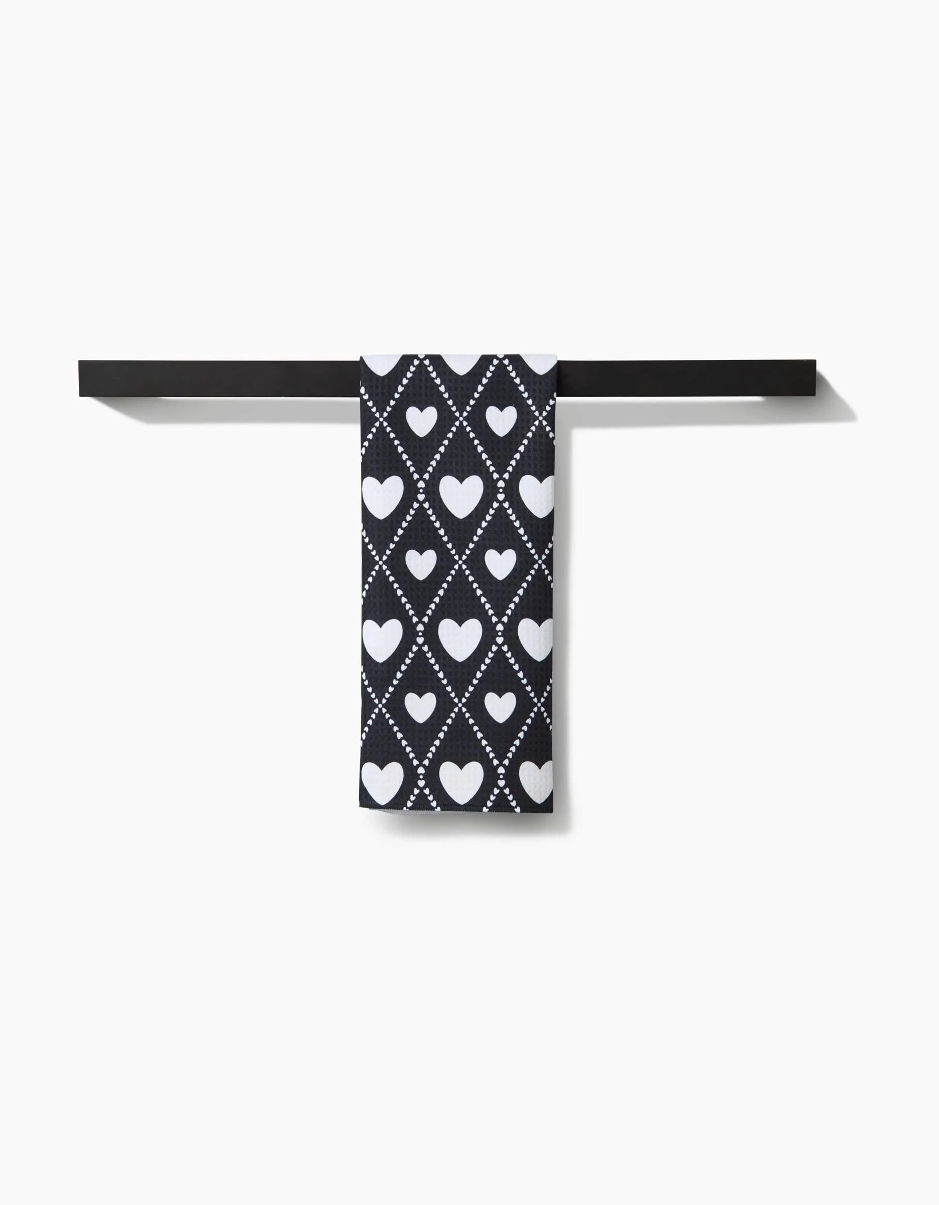 The Onyx Love Lattice hand towel by Charlie Rowan Designs, made from recycled materials, displays a black design with white heart and diamond patterns and hangs on a black towel bar against a white background.